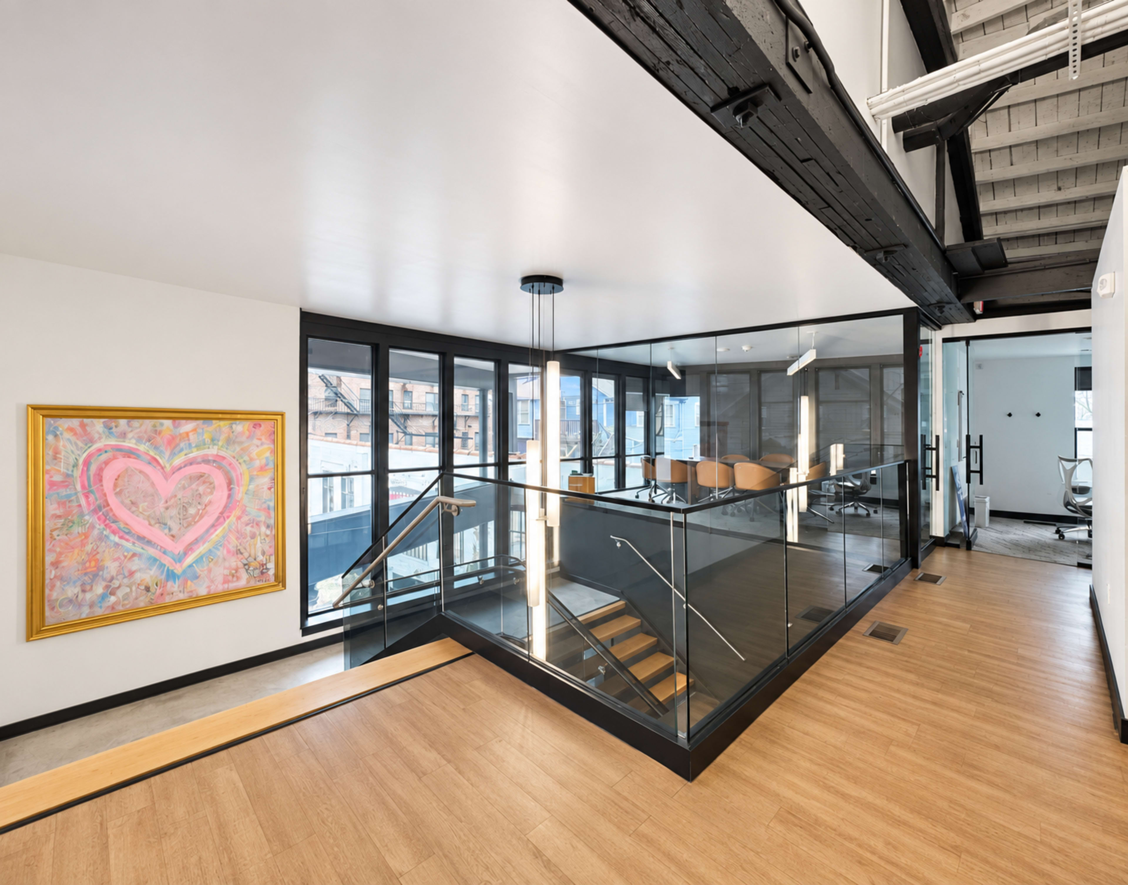 The image shows a modern office space with large windows, a glass railing staircase, and an adjacent conference room featuring a colorful heart painting on the wall.