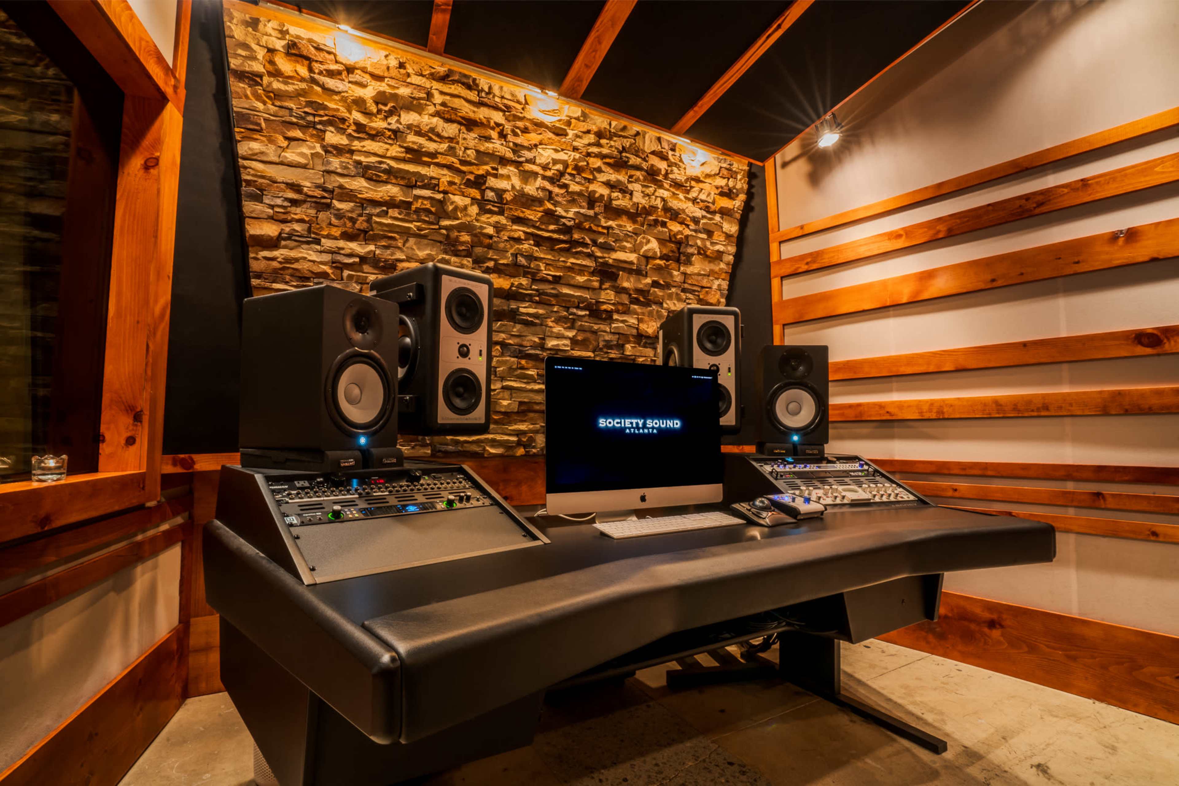 A modern recording studio with a stone accent wall, a mixing desk, two large speakers, and a computer displaying "SOCIETY SOUND."