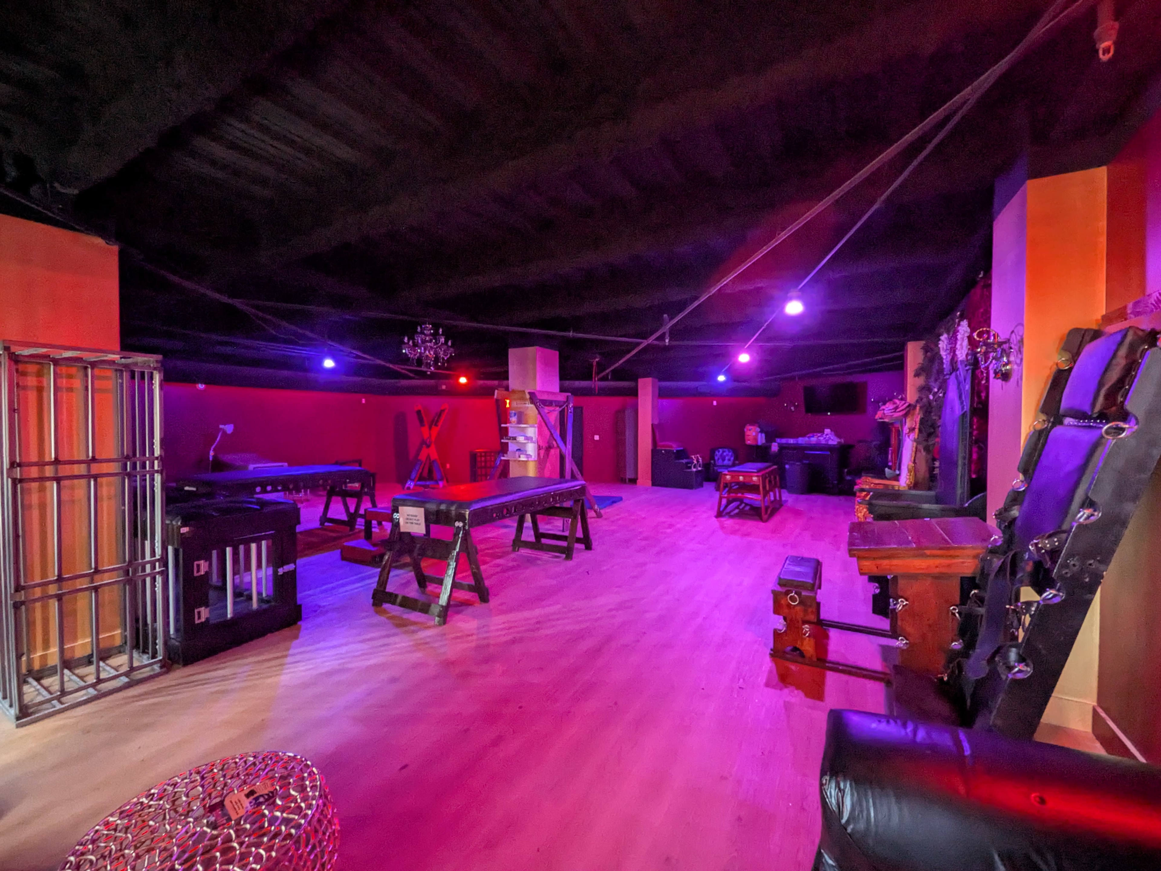 The image shows a dimly lit room designed for various activities, featuring multiple platforms, bondage equipment, and colored lighting.