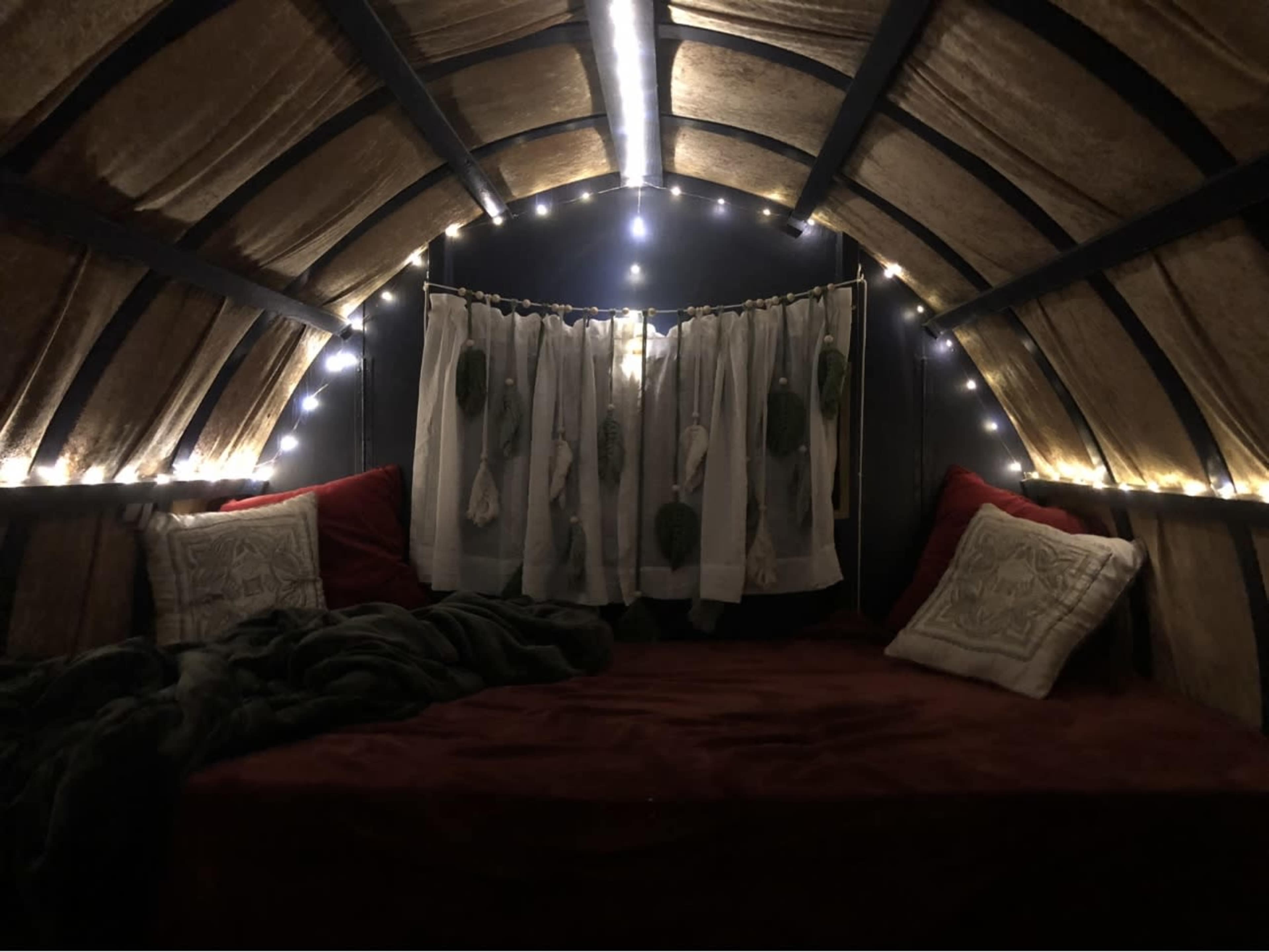 The image shows a cozy, dimly lit interior space with a bed covered in a red blanket, surrounded by soft lighting and sheer curtains.