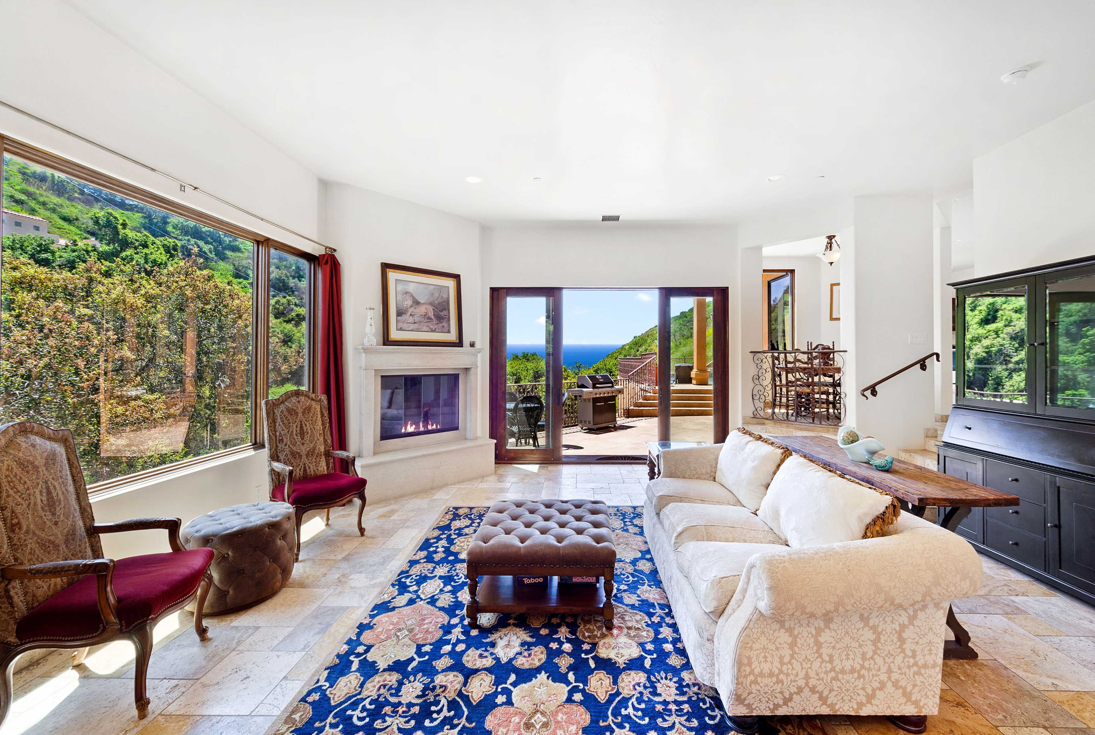 A sunny living room features a white sofa, two armchairs, a coffee table, and large windows with a view of the ocean.