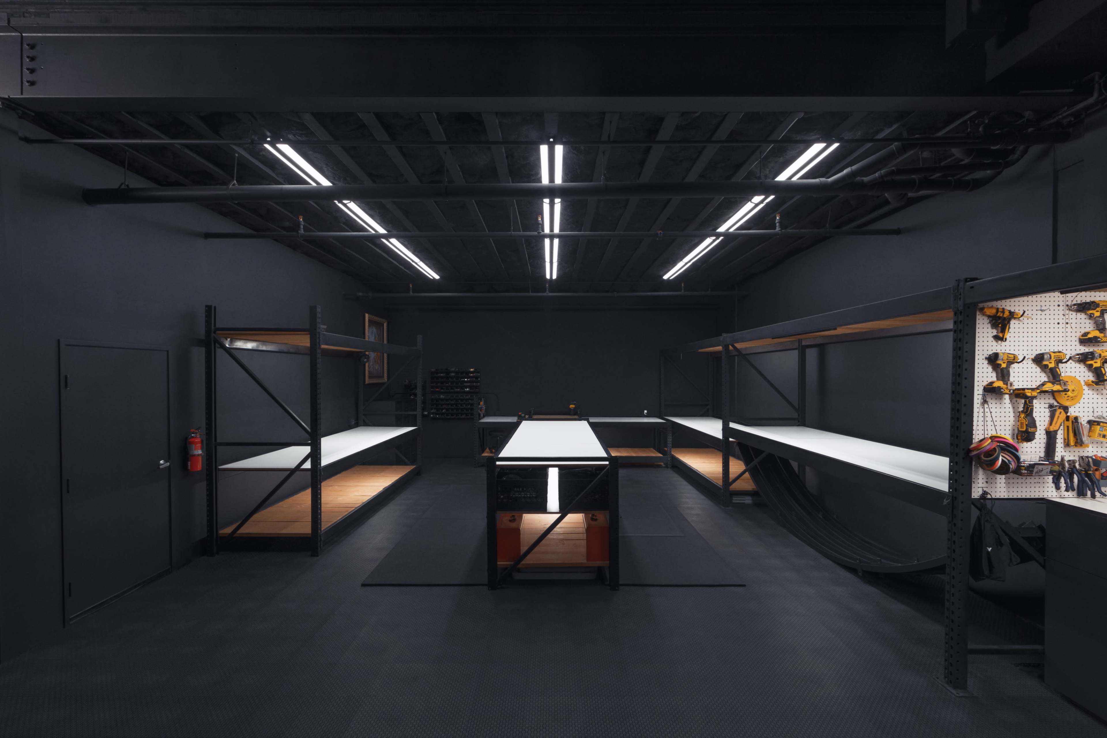 A dimly lit workshop with black walls, illuminated by overhead lights, featuring industrial shelving and a central workbench.