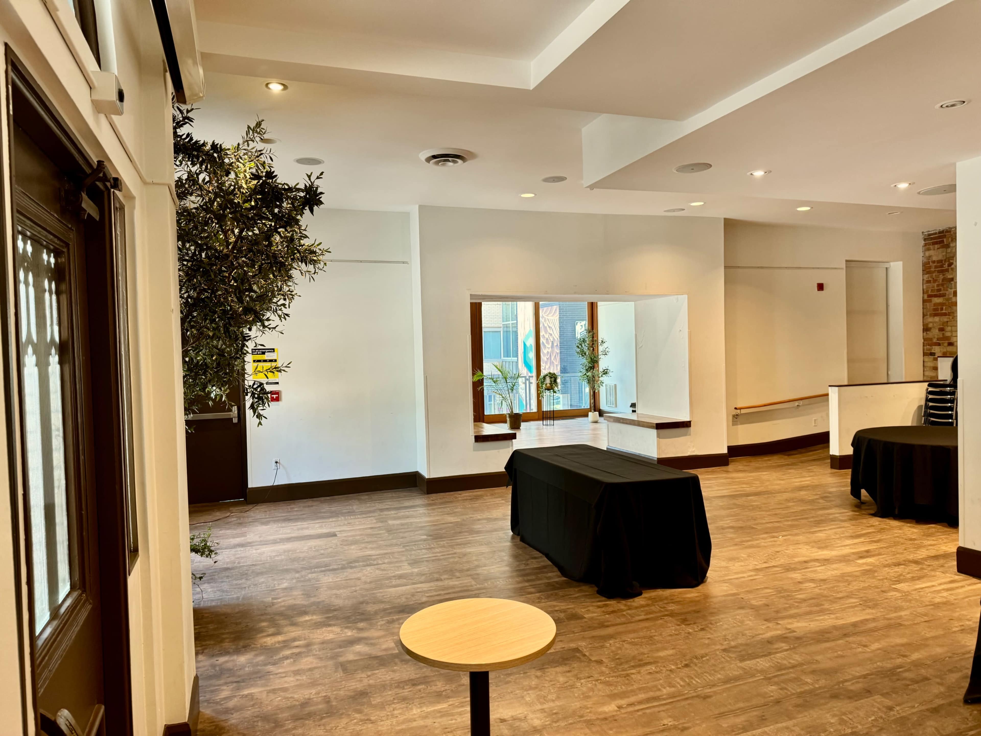 The image shows a spacious indoor area with wooden flooring, two black-covered tables, and a potted plant near a large window.