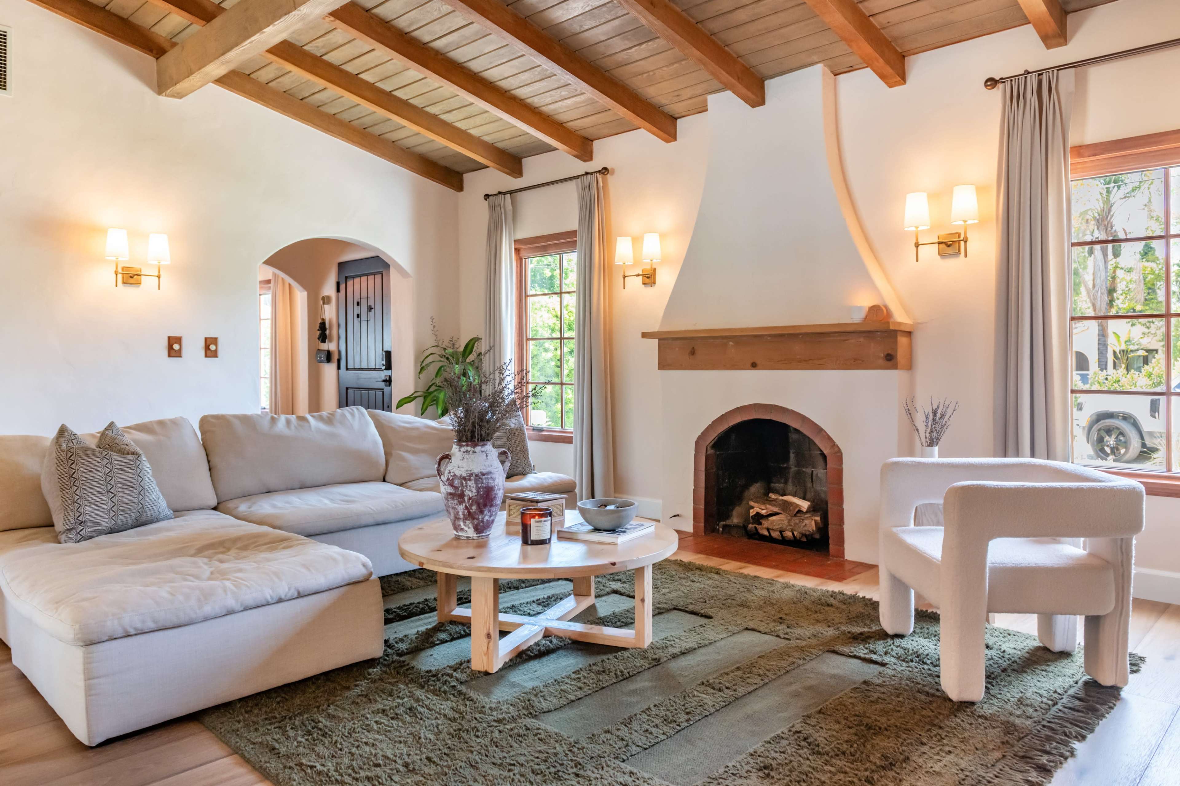 The image shows a cozy living room with a sectional sofa, a round coffee table, and a fireplace, surrounded by large windows that let in natural light.