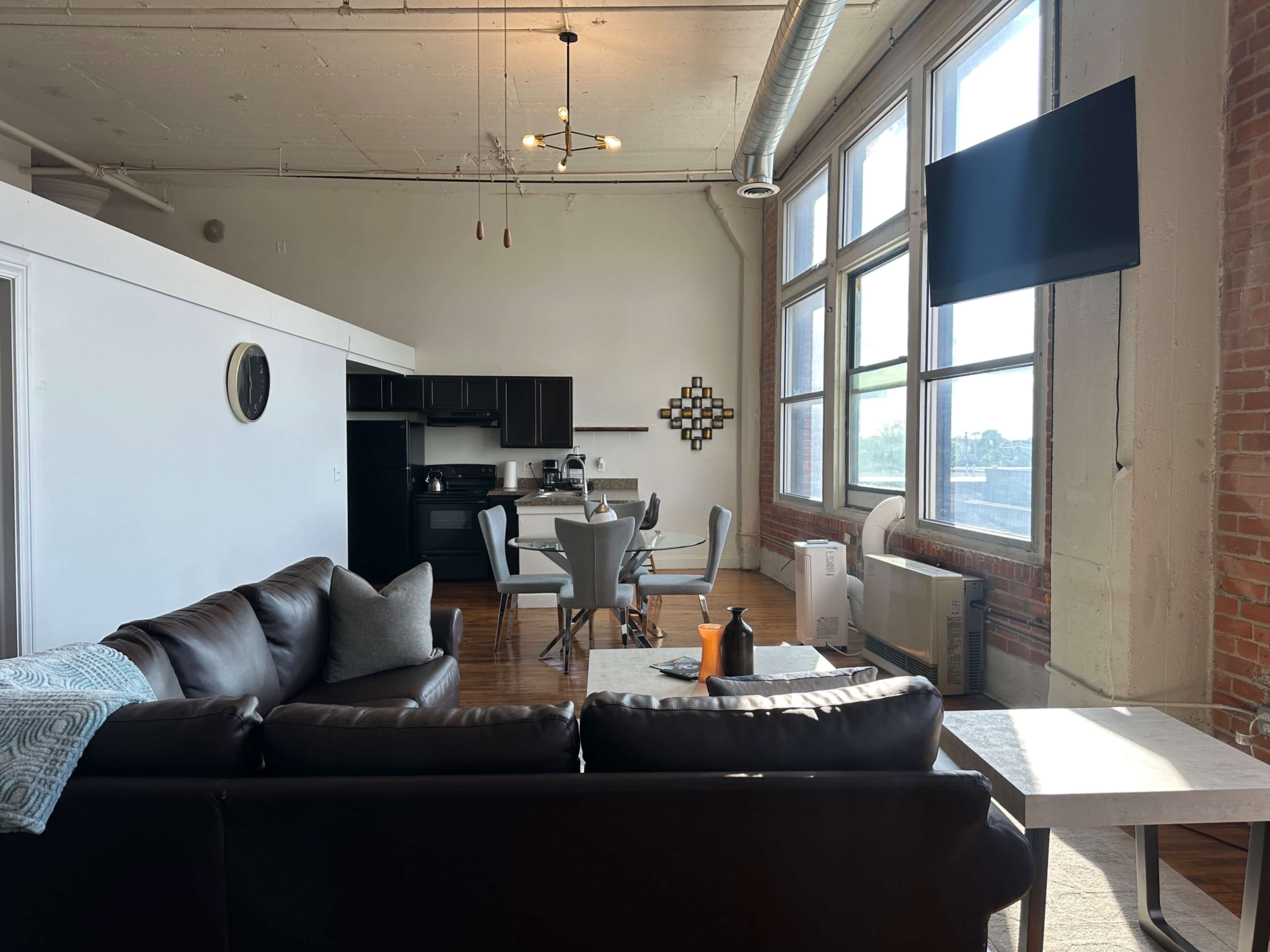 The image shows a modern apartment interior featuring a living area with a dark sofa, a dining table, and large windows that let in natural light.