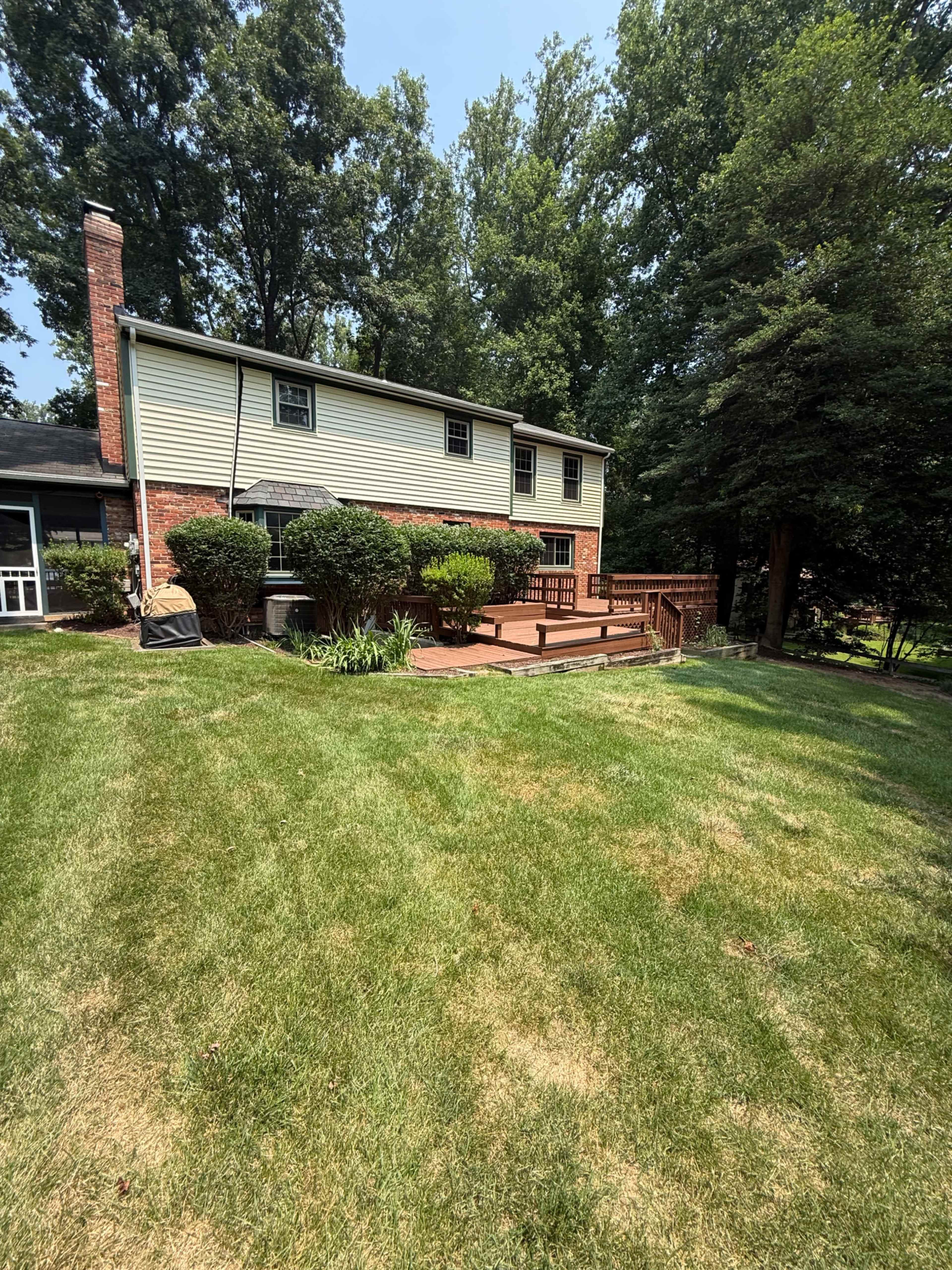 The image shows a two-story house with a wooden deck in the backyard surrounded by well-maintained grass and trees.