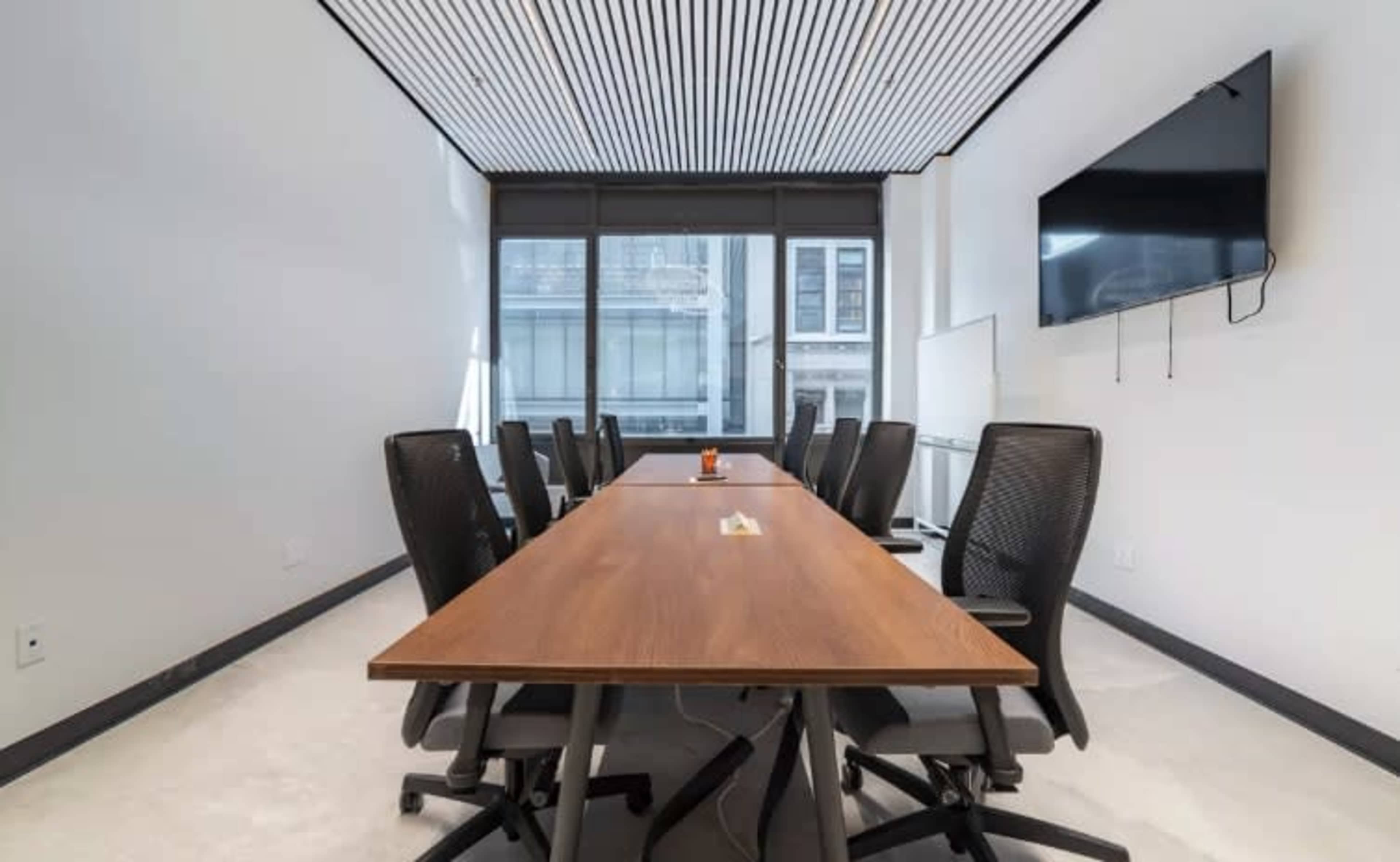 A modern conference room features a long wooden table surrounded by black mesh chairs, with large windows and a wall-mounted TV.