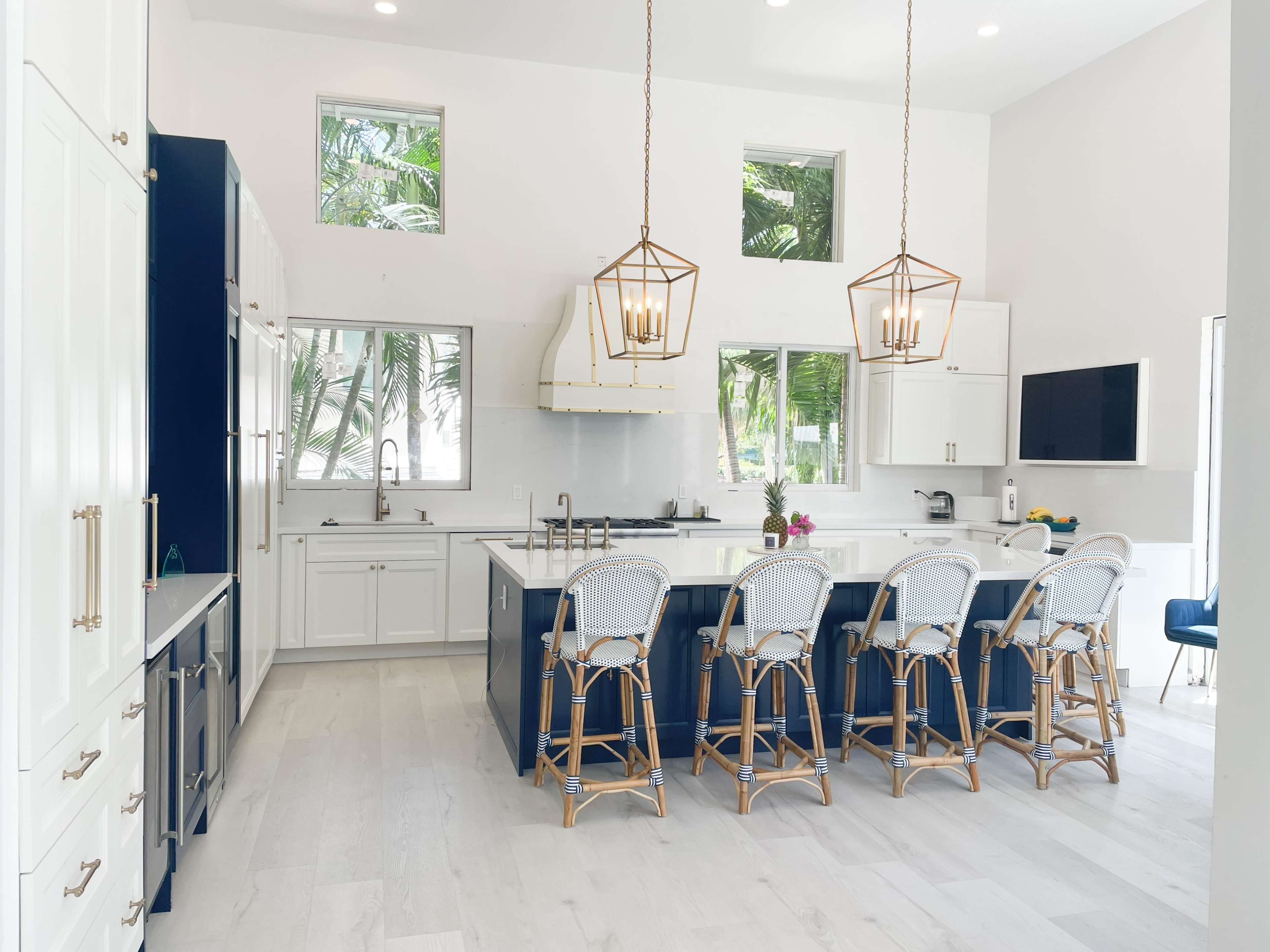 A modern kitchen features a large island with six barstools, white cabinets, stainless steel appliances, and large windows overlooking tropical greenery.