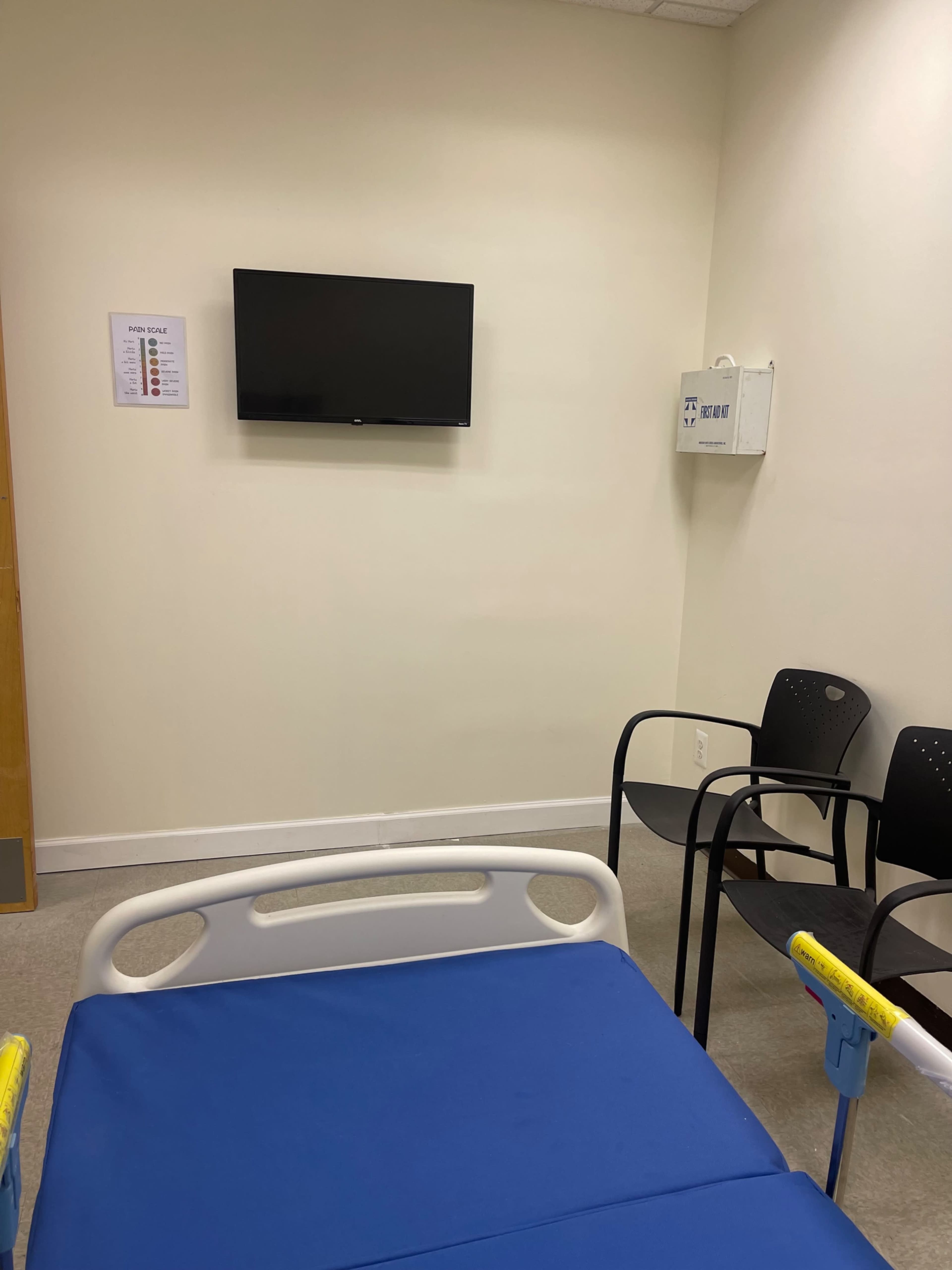 The image shows a hospital room with a bed featuring a blue mattress, a wall-mounted television, and a few black chairs positioned against the wall.