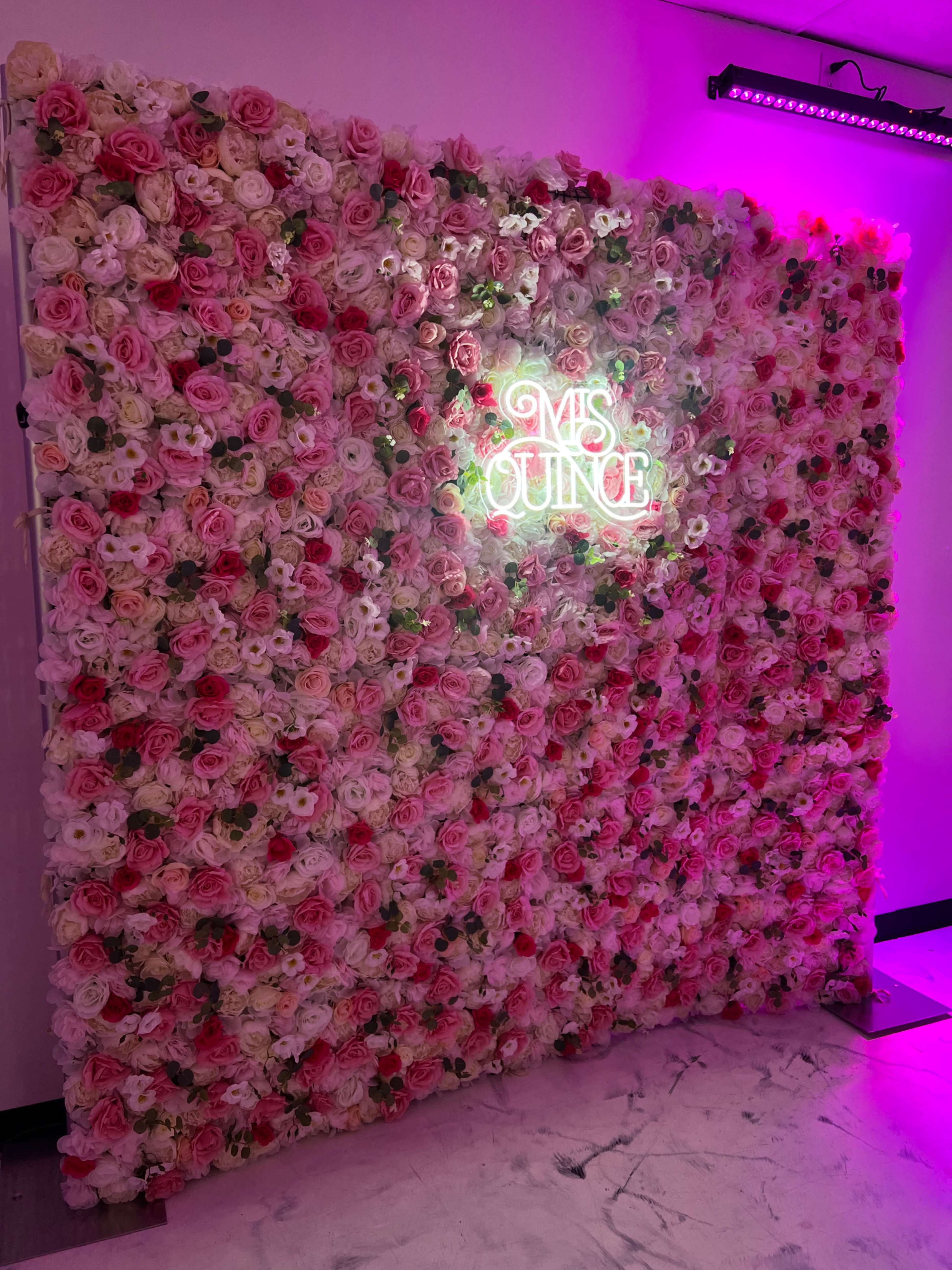 The image shows a decorative wall covered with faux flowers in various shades of pink and white, featuring a neon sign that reads "Ms. Quince."
