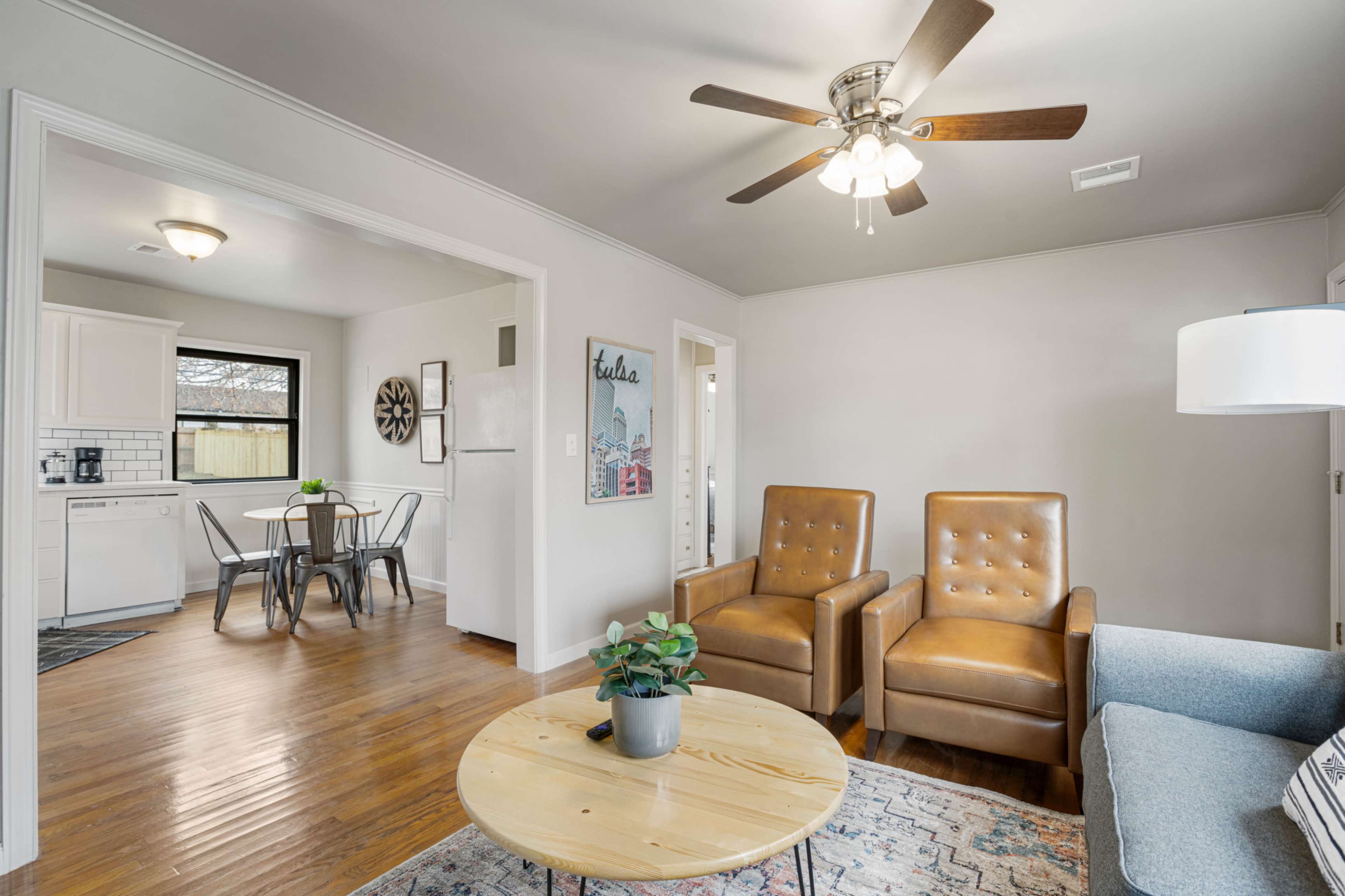 The image shows a living room with two brown armchairs, a round wooden coffee table, and a light blue couch, leading into a dining area with a table and chairs.