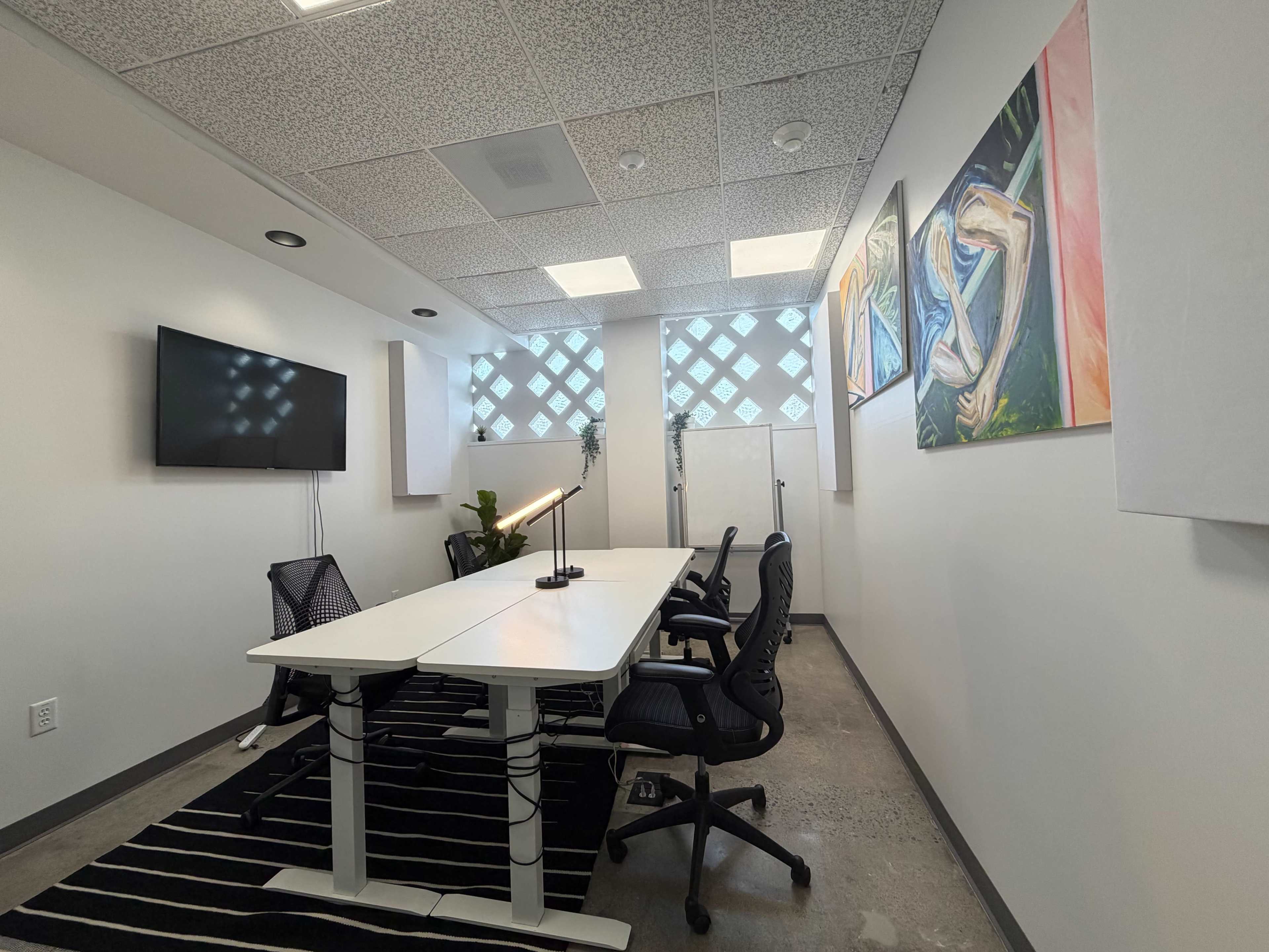The image shows a modern conference room with a long table, black chairs, a wall-mounted TV, and abstract artwork on the walls.
