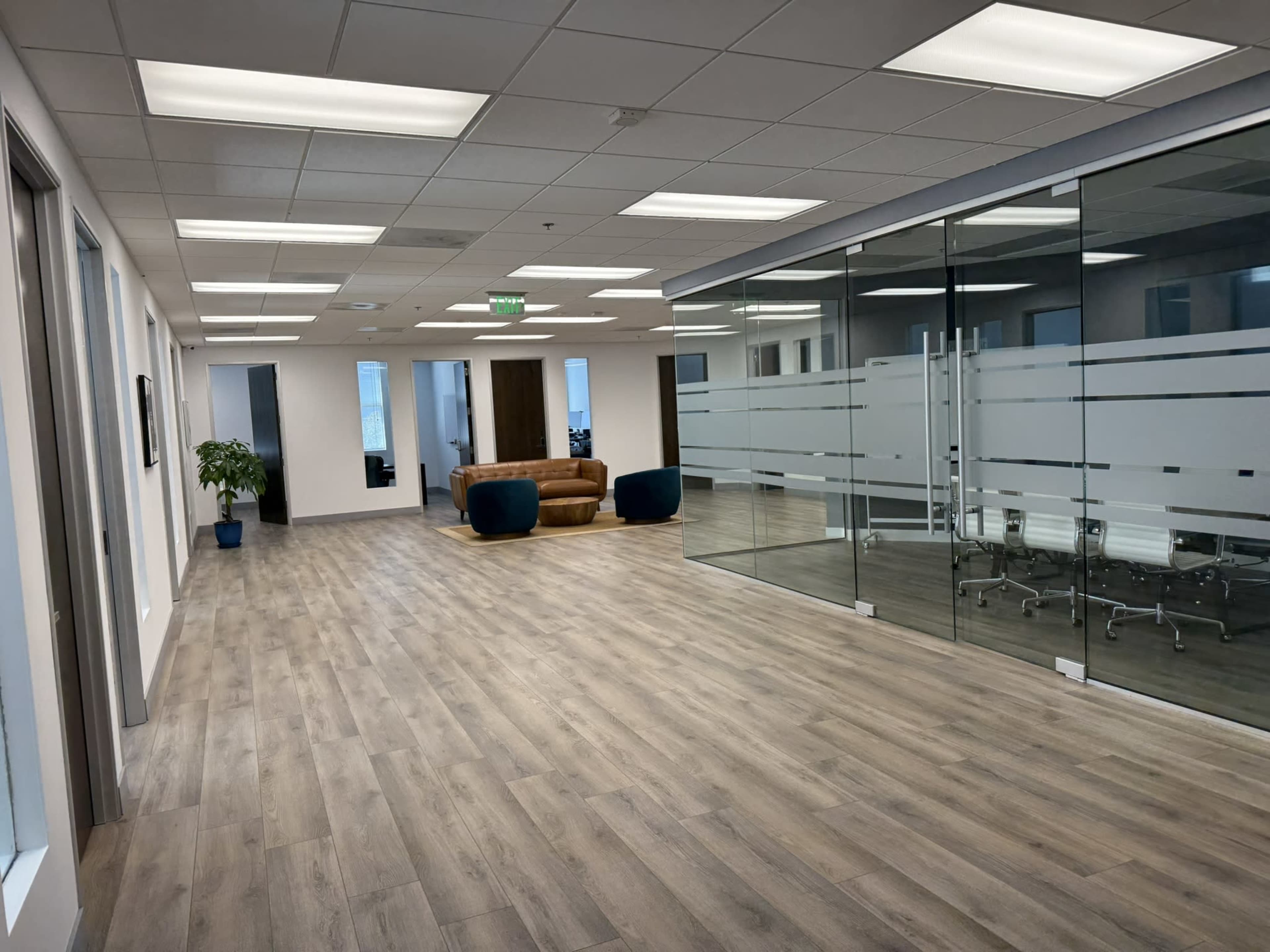 The image shows a modern office interior featuring a spacious hallway with wooden floors, a lounge area with a brown sofa, and a glass-walled meeting room.