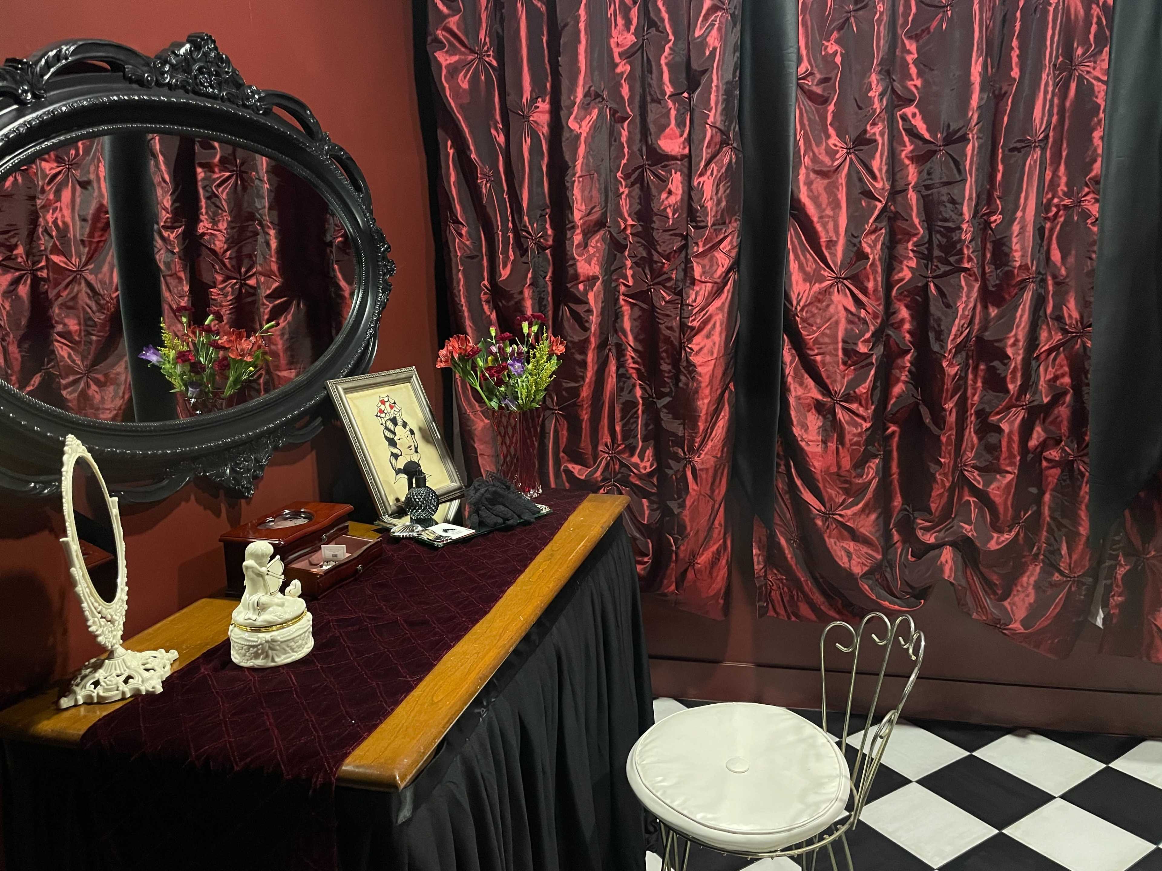 The scene features a table adorned with a decorative mirror, a framed photo, and flowers, set against deep red curtains and a black-and-white checkered floor.