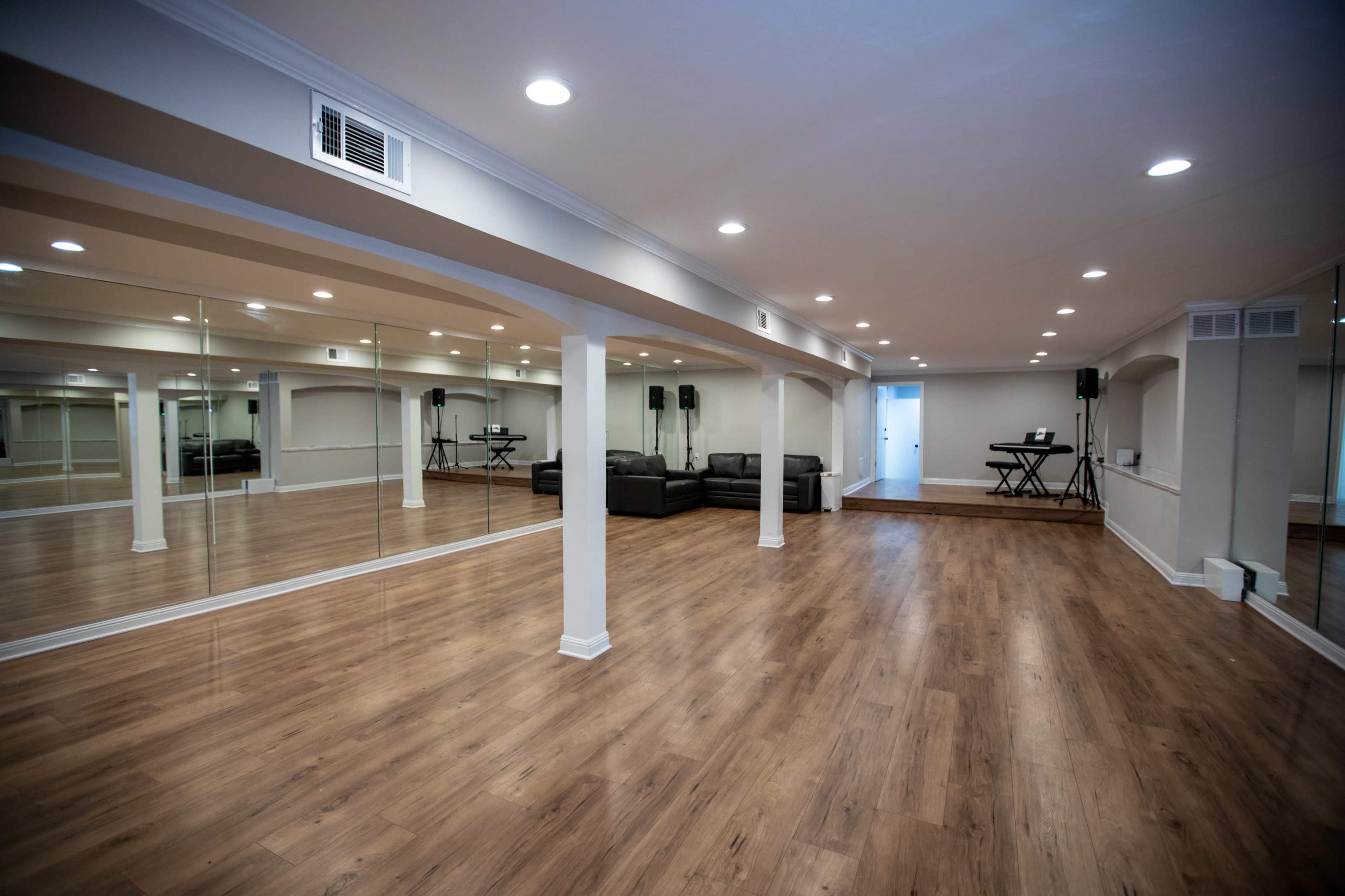 The image shows a spacious, well-lit room with large mirrors on one wall, hardwood flooring, and a seating area featuring a dark sofa.