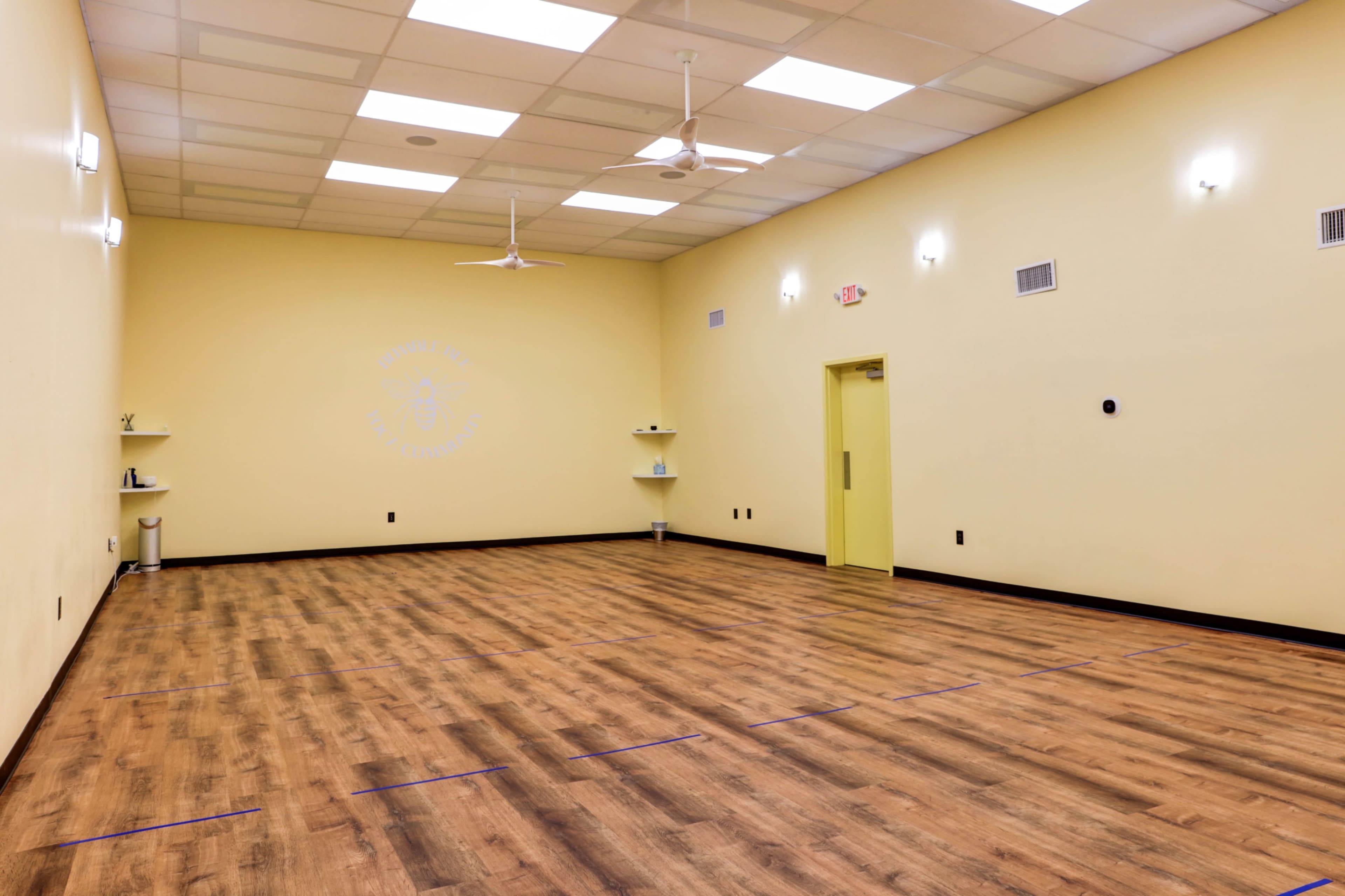 An empty, well-lit room with yellow walls and a wooden floor, featuring ceiling fans and minimalist decor.