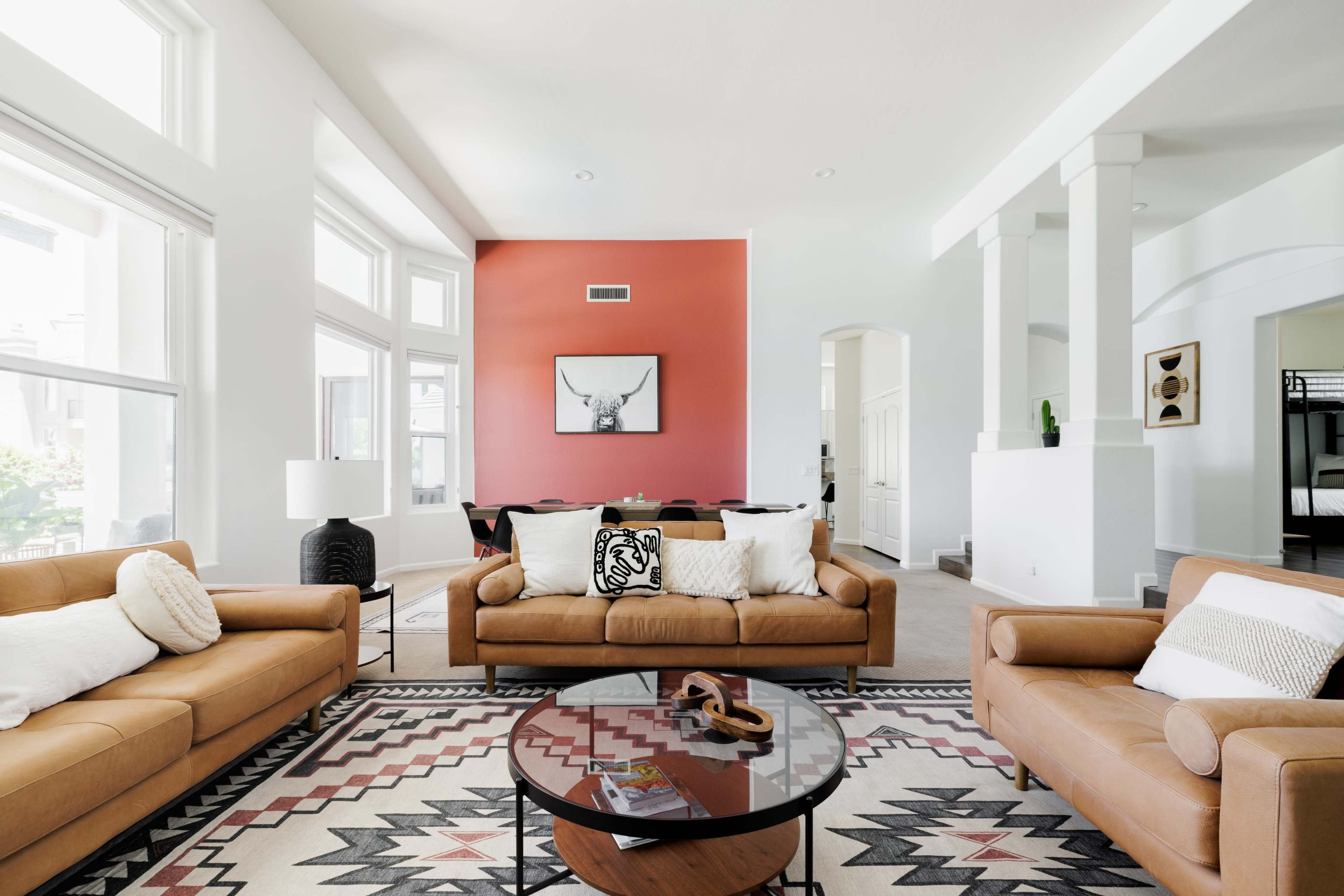 The living room features two brown leather sofas facing a round coffee table on a patterned rug, with large windows allowing natural light and a red accent wall displaying an artwork.