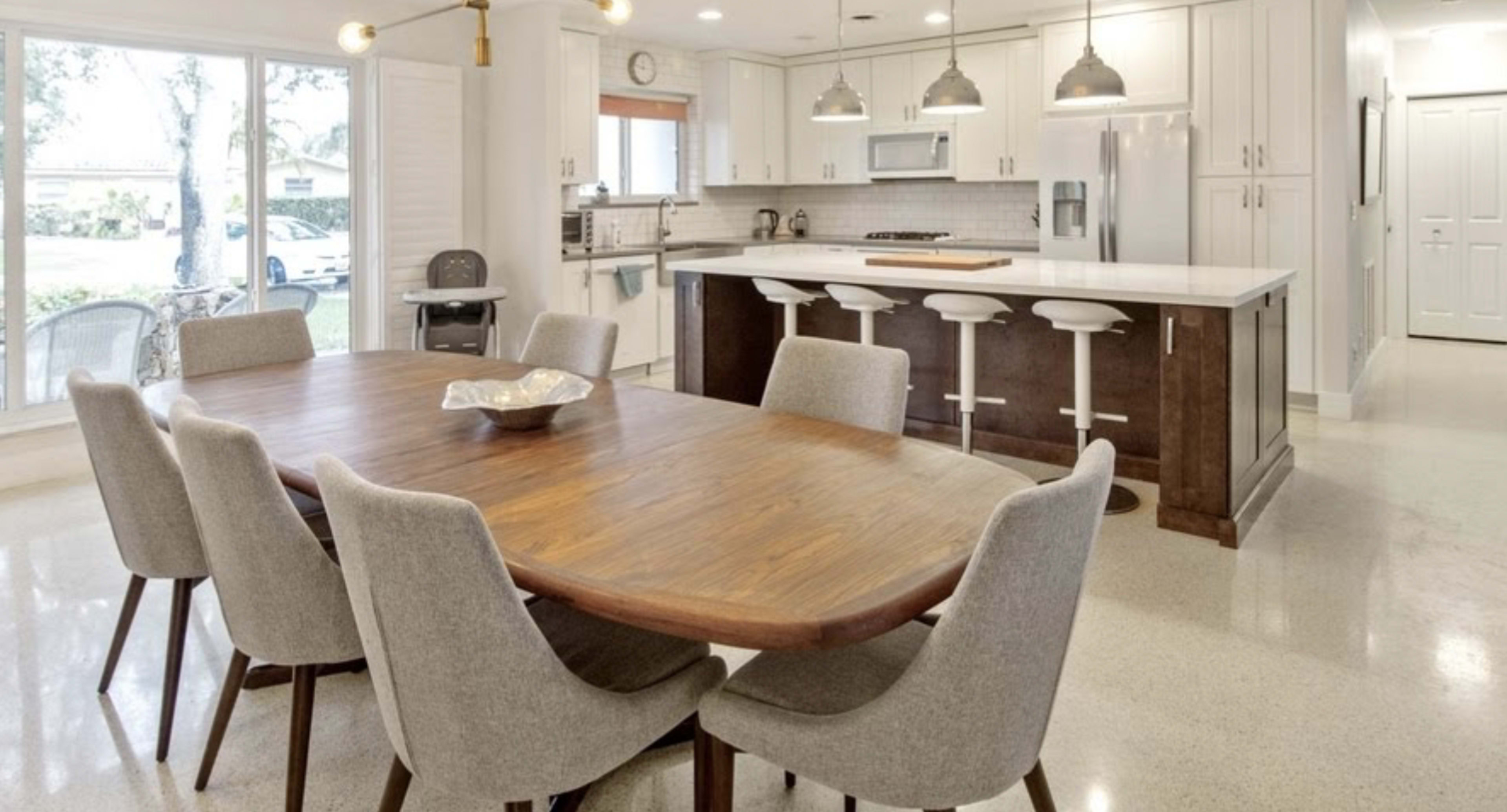 The image shows a modern kitchen and dining area featuring a wooden dining table surrounded by grey upholstered chairs, with a kitchen island and pendant lighting above.