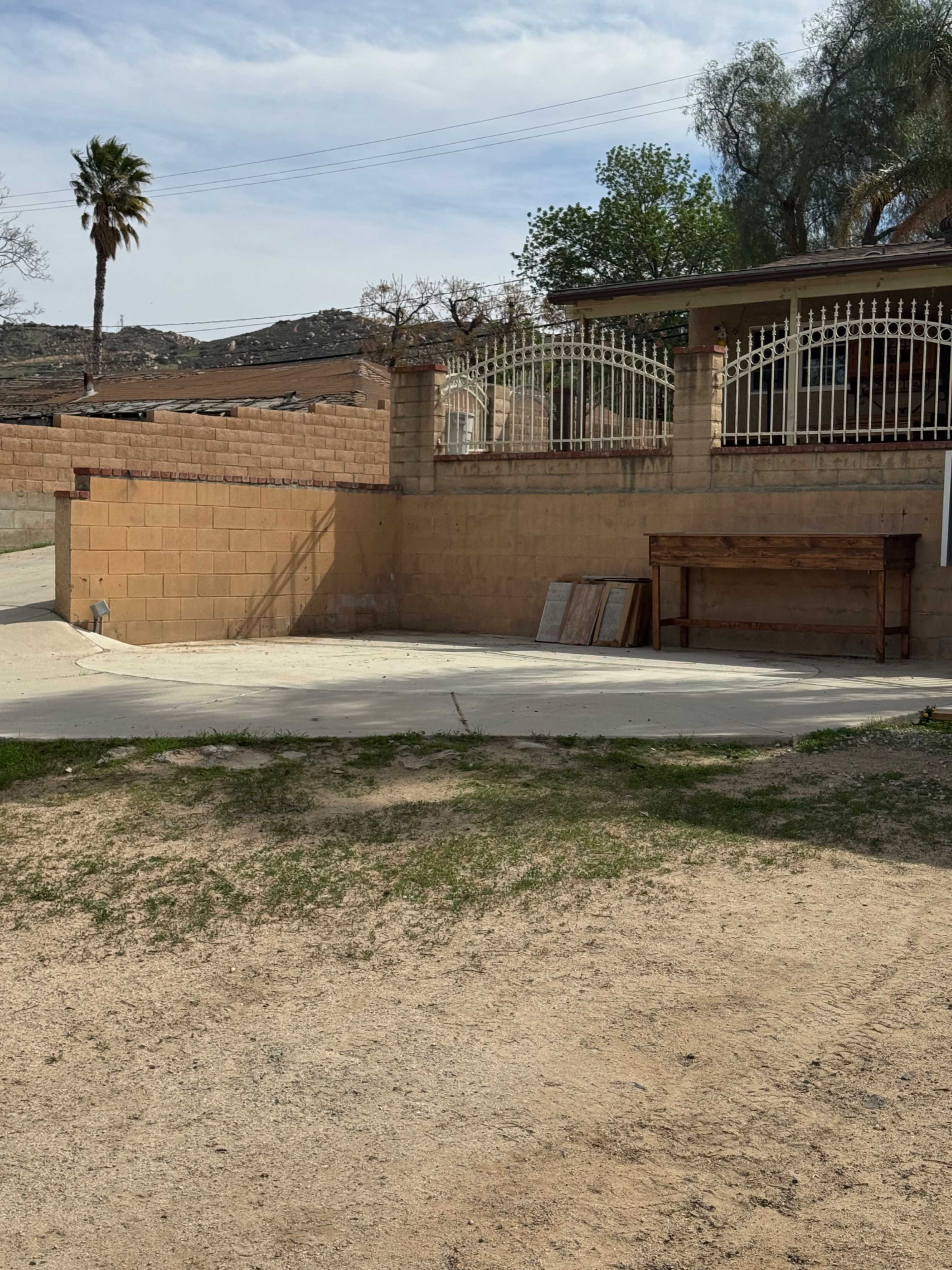 The image shows a dirt area in front of a low brick wall with a wooden table and palm tree in the background.