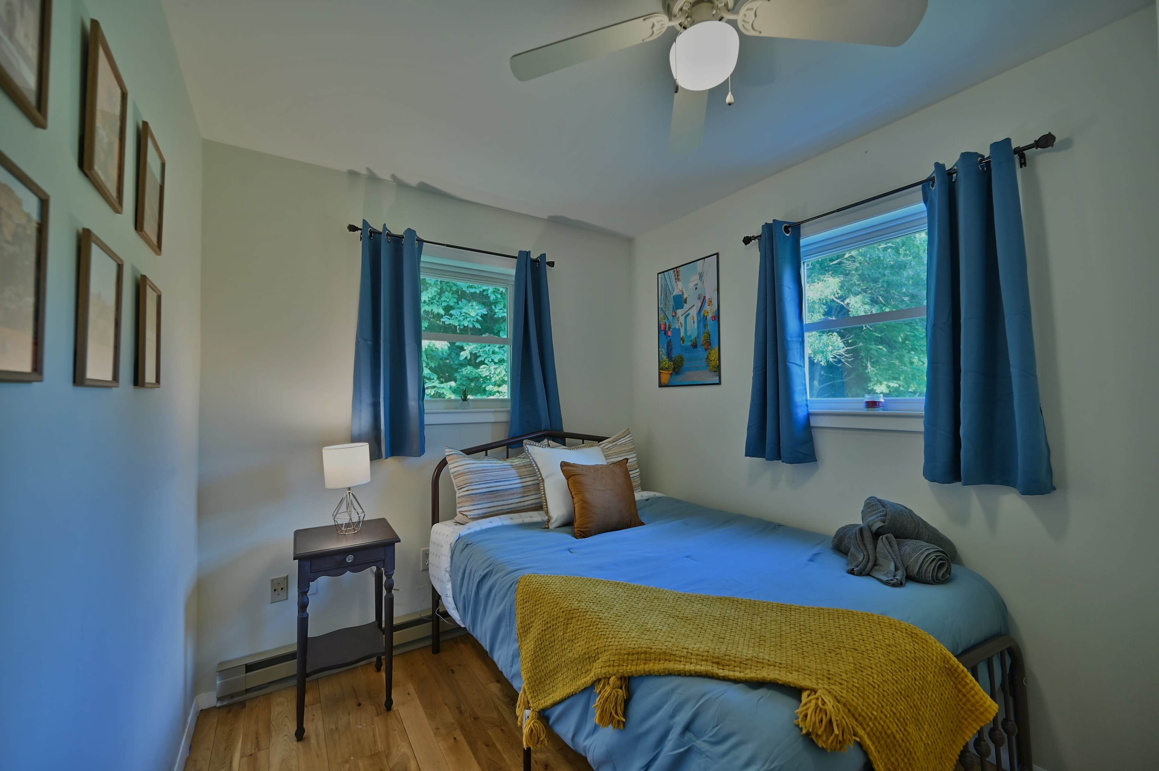 The image shows a small bedroom with a bed, bedside table, and two windows, adorned with blue curtains and decorative wall art.