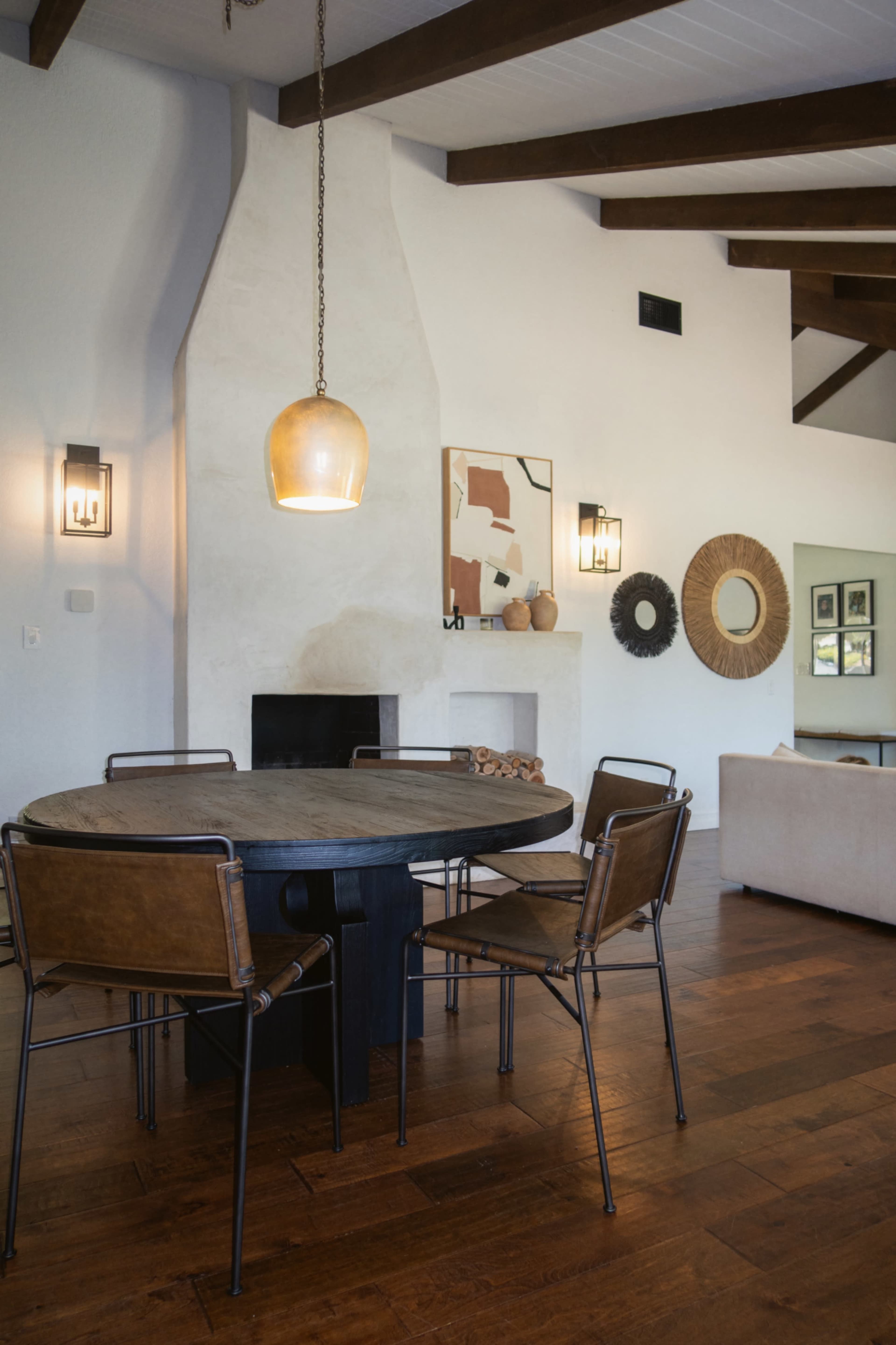The image shows a modern dining area featuring a round wooden table surrounded by metal and leather chairs, with a fireplace and wall art in the background.