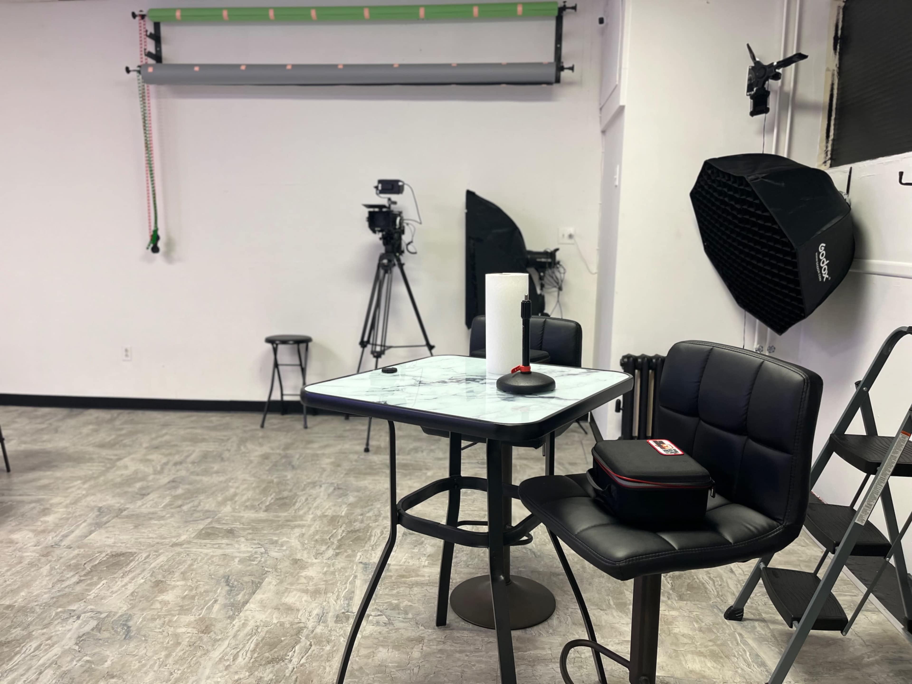 A studio space featuring a table with a microphone, chairs, and video recording equipment positioned in the background.
