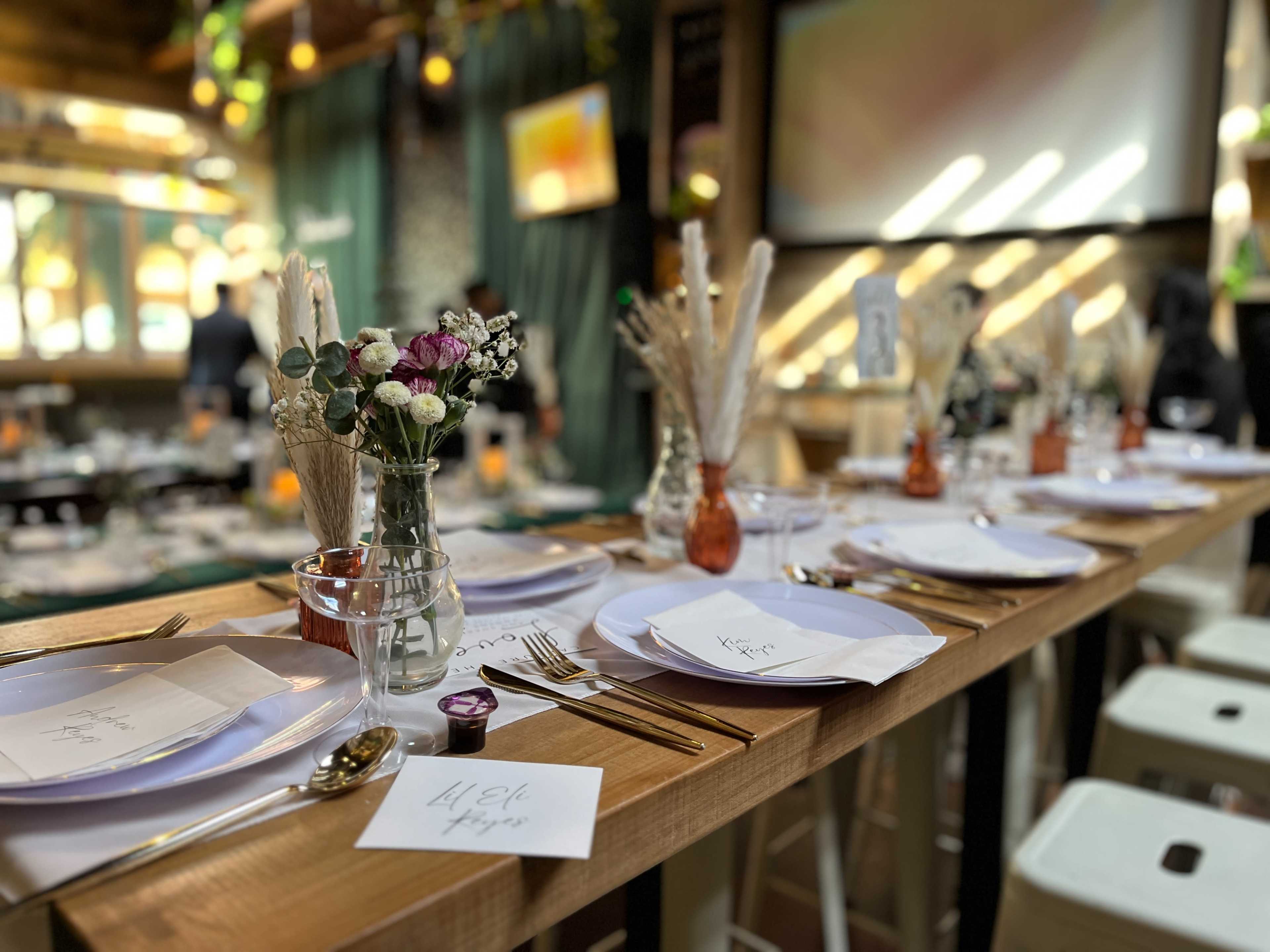 A long dining table is set with white plates, cutlery, and small floral arrangements, ready for guests in a well-lit venue.