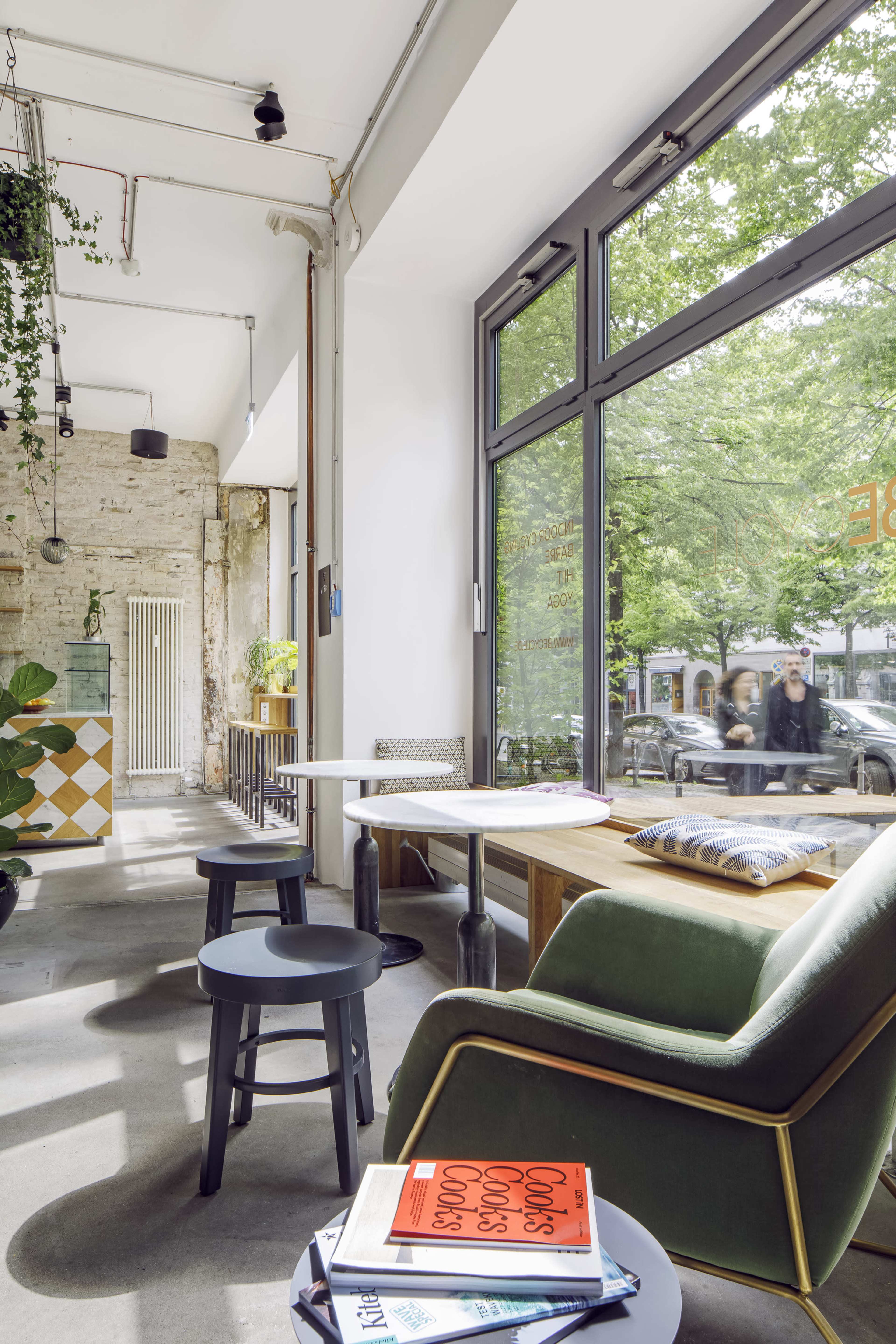 A modern café interior features a mix of green seating, wooden tables, and large windows overlooking a tree-lined street.