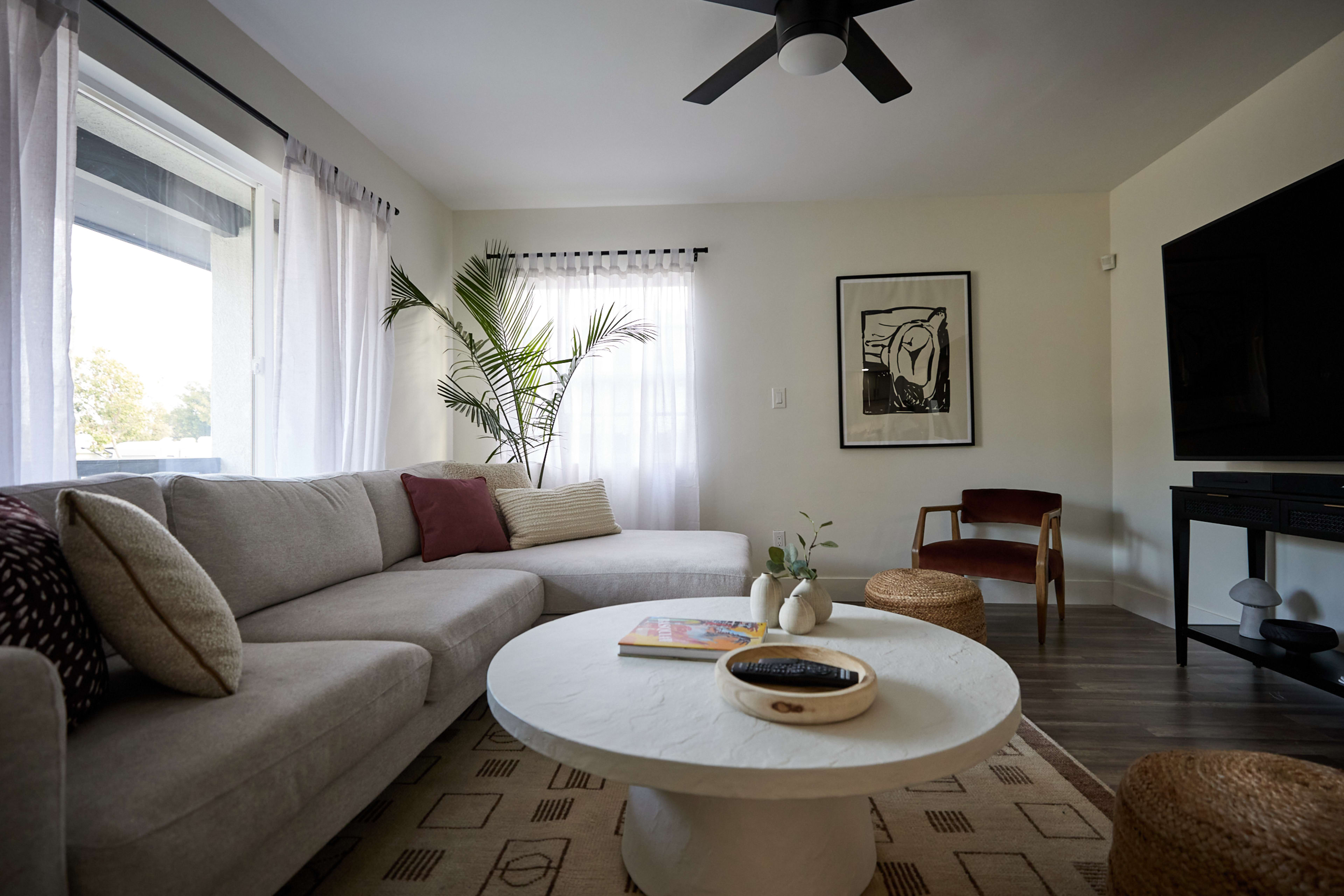 The image shows a modern living room featuring a light gray sectional sofa, a round coffee table, and a plant near a window with sheer curtains.