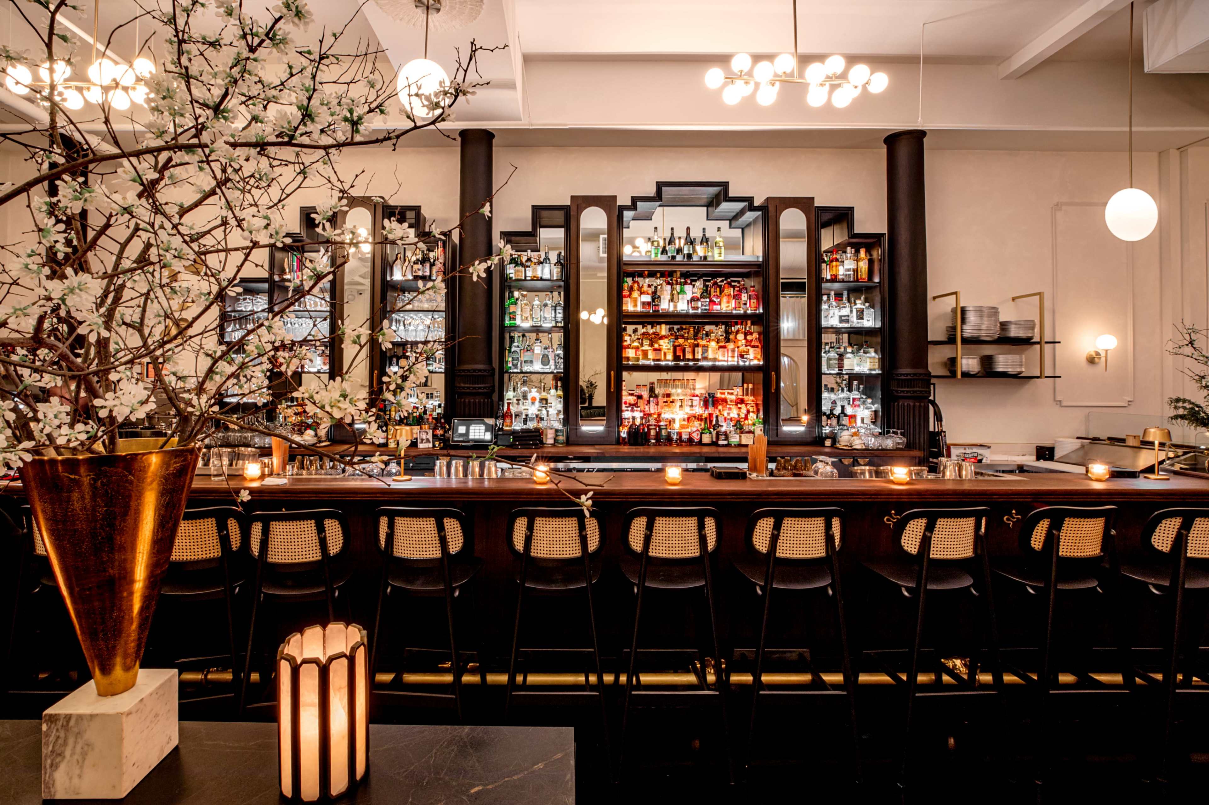 The image depicts a stylish bar interior with a long counter, shelves filled with various bottles, decorative lighting, and floral arrangements.