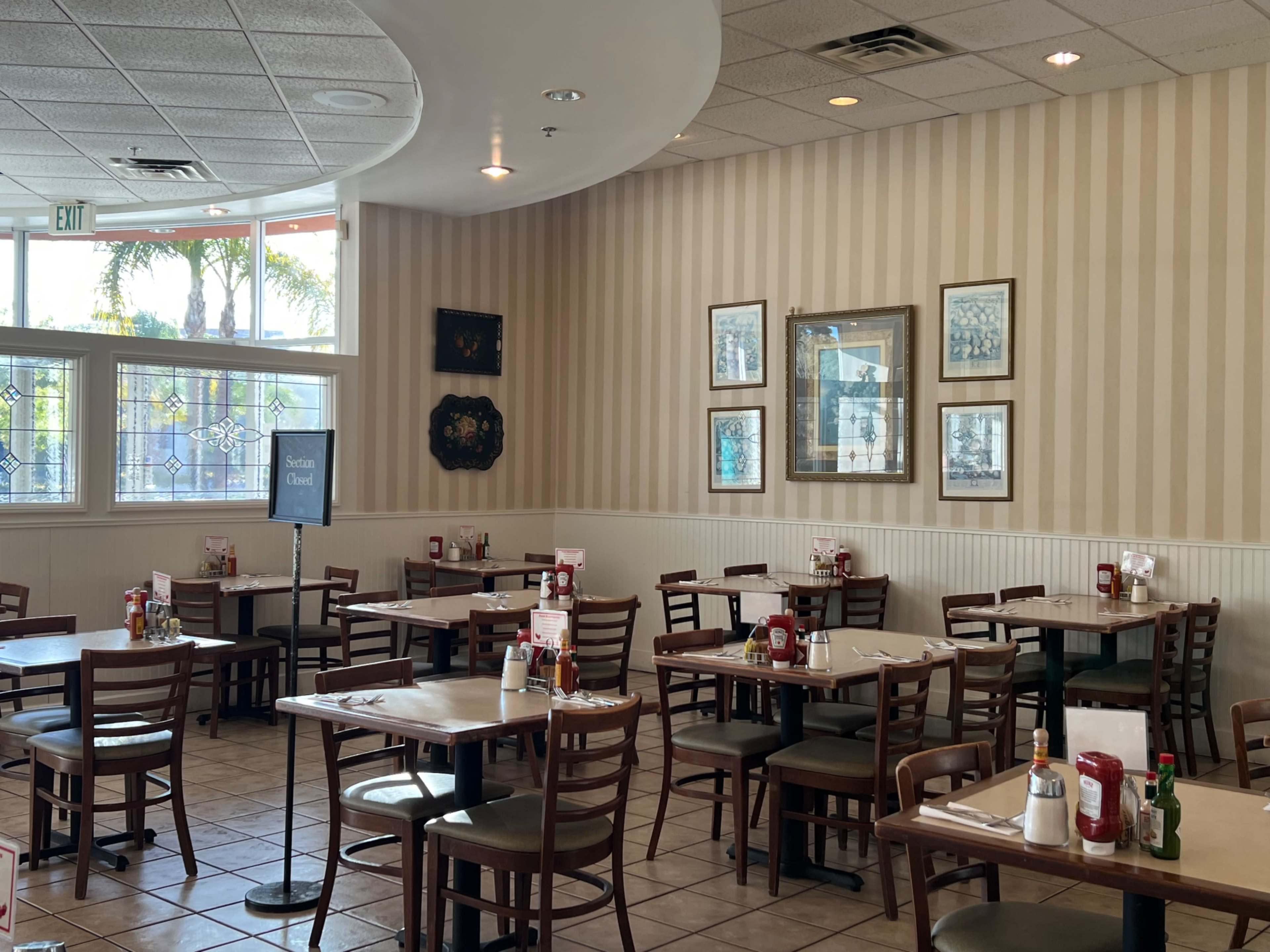 The image shows a spacious restaurant interior with several empty tables and a sign indicating the section is closed, framed by striped wallpaper and adorned with framed pictures on the walls.