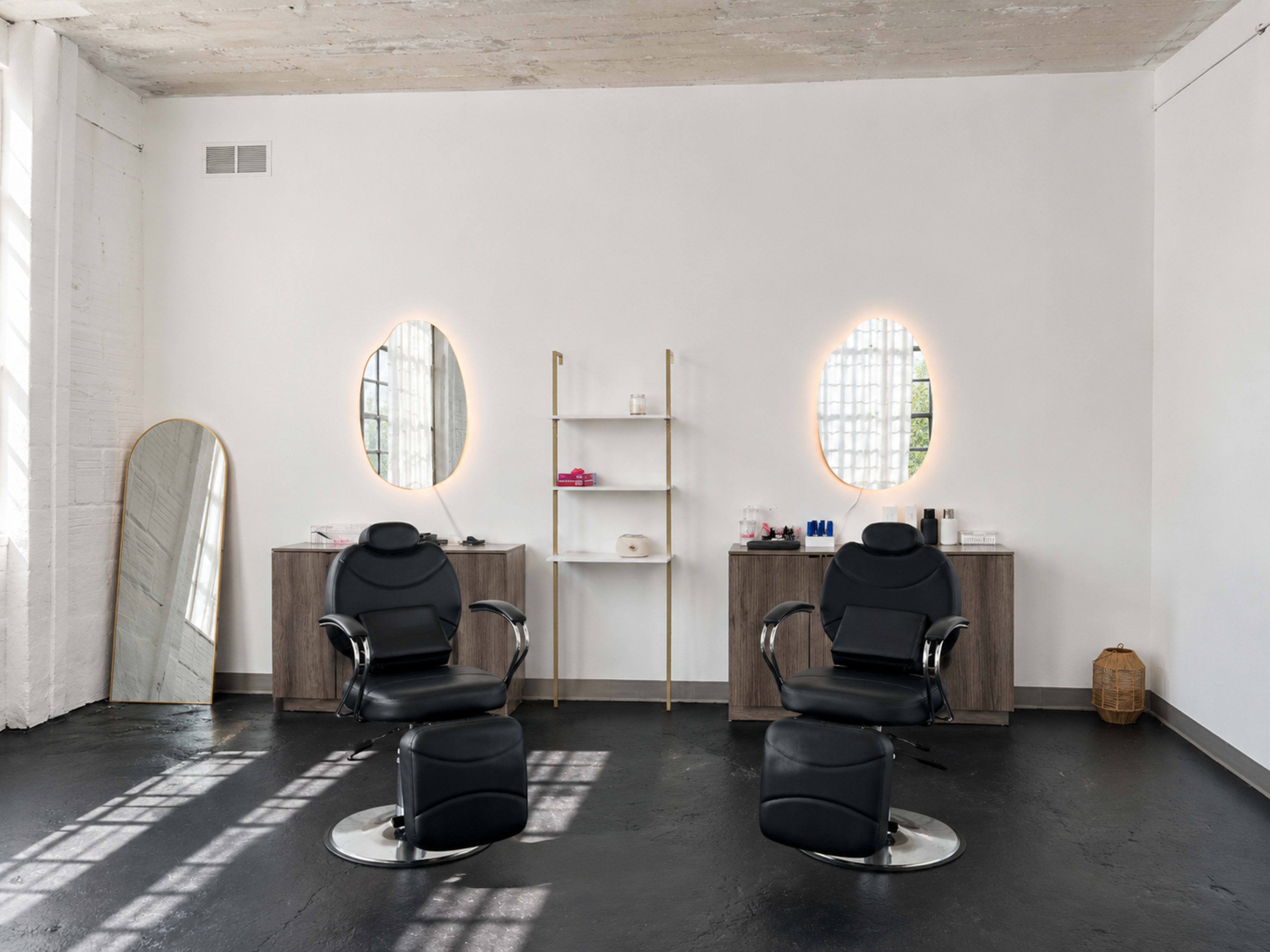 The image shows a modern hair salon with two black styling chairs, a large mirror, minimalist shelving, and natural light streaming through windows.