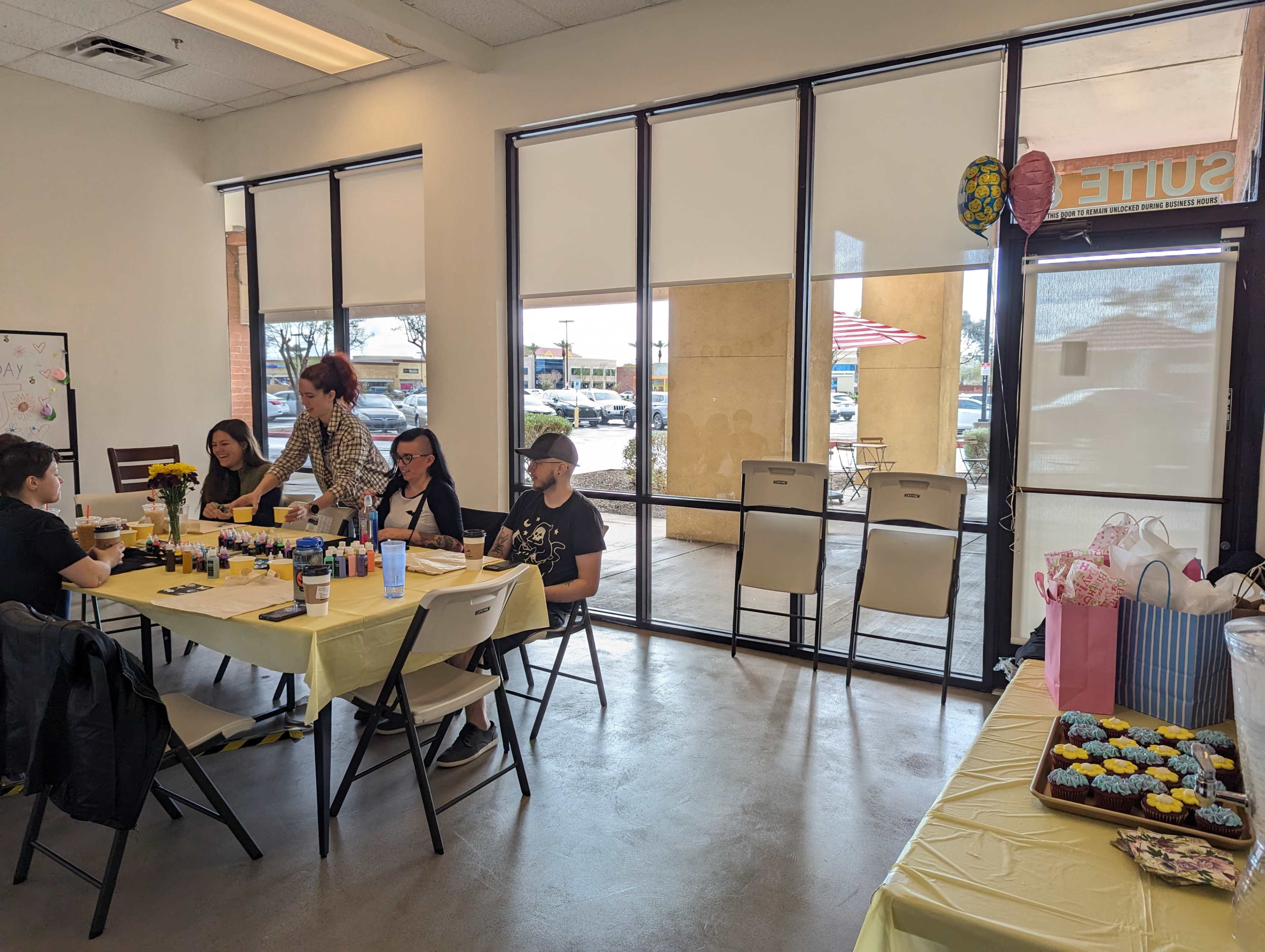 A group of people sits at a table covered with craft supplies in a brightly lit room with large windows.