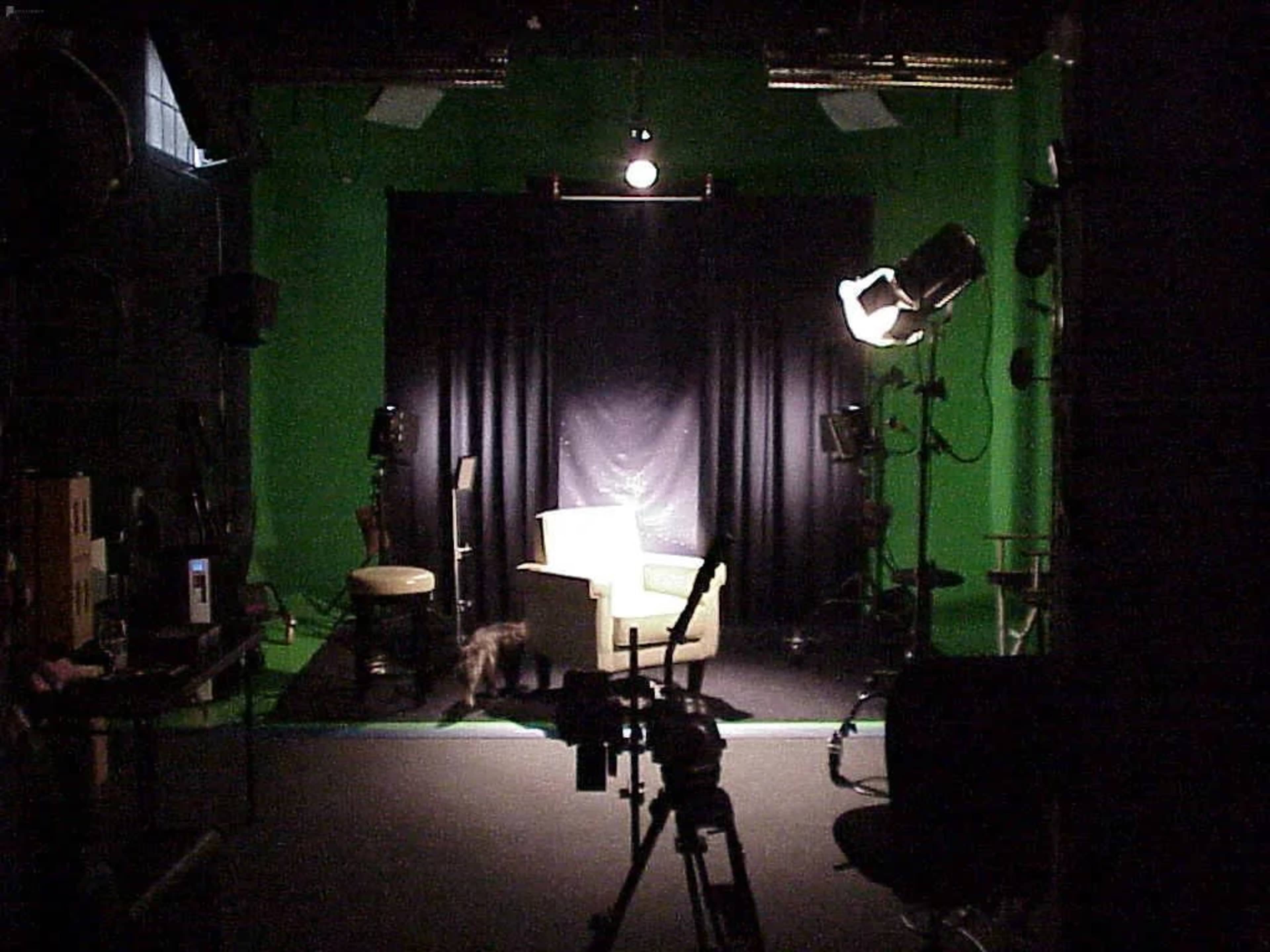 The image shows a dimly lit studio with a green backdrop, a spotlight focused on a chair, and various lighting and camera equipment positioned around the scene.