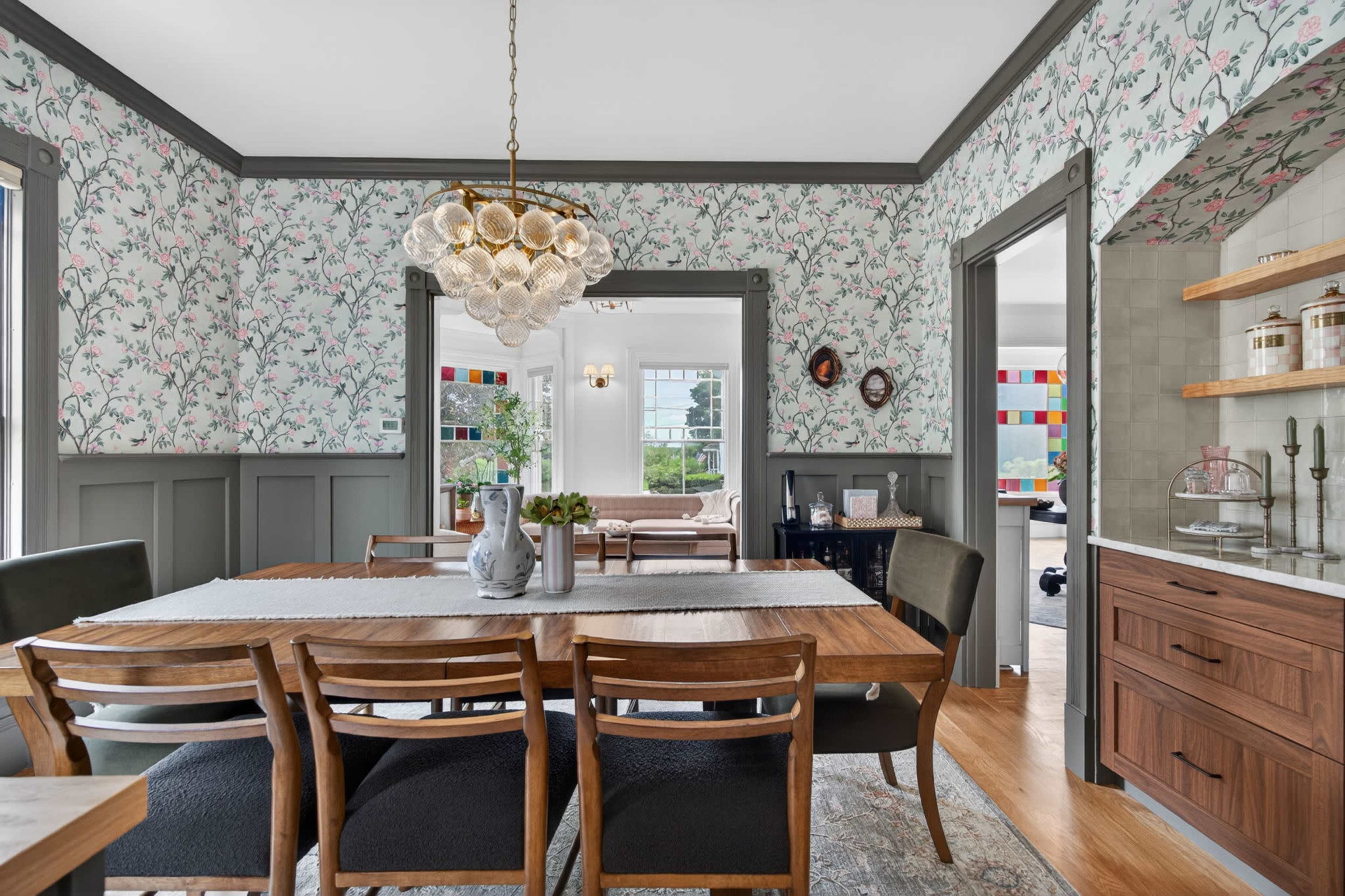 A dining room features a long wooden table surrounded by chairs, with floral wallpaper and a chandelier hanging above.
