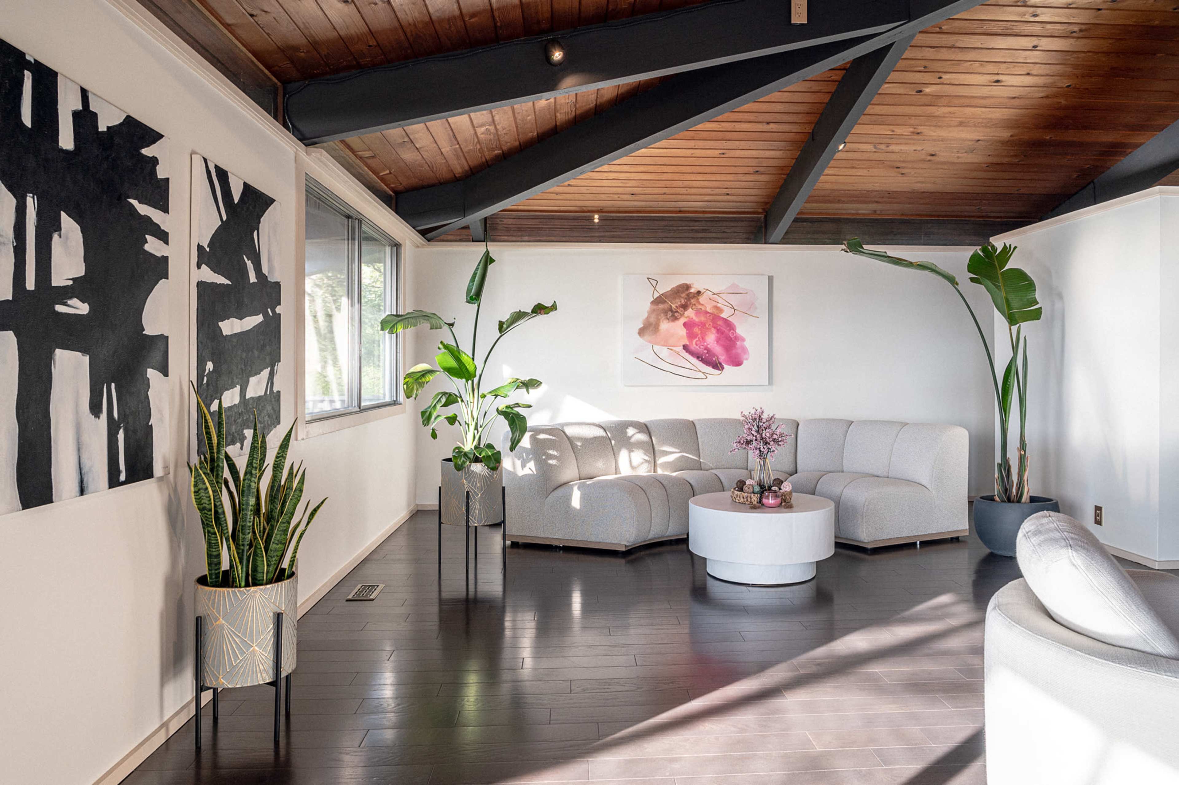 A modern living room with a circular coffee table, light-colored seating, and large plants placed in decorative pots against a backdrop of vaulted wooden beams and abstract artwork on the walls.