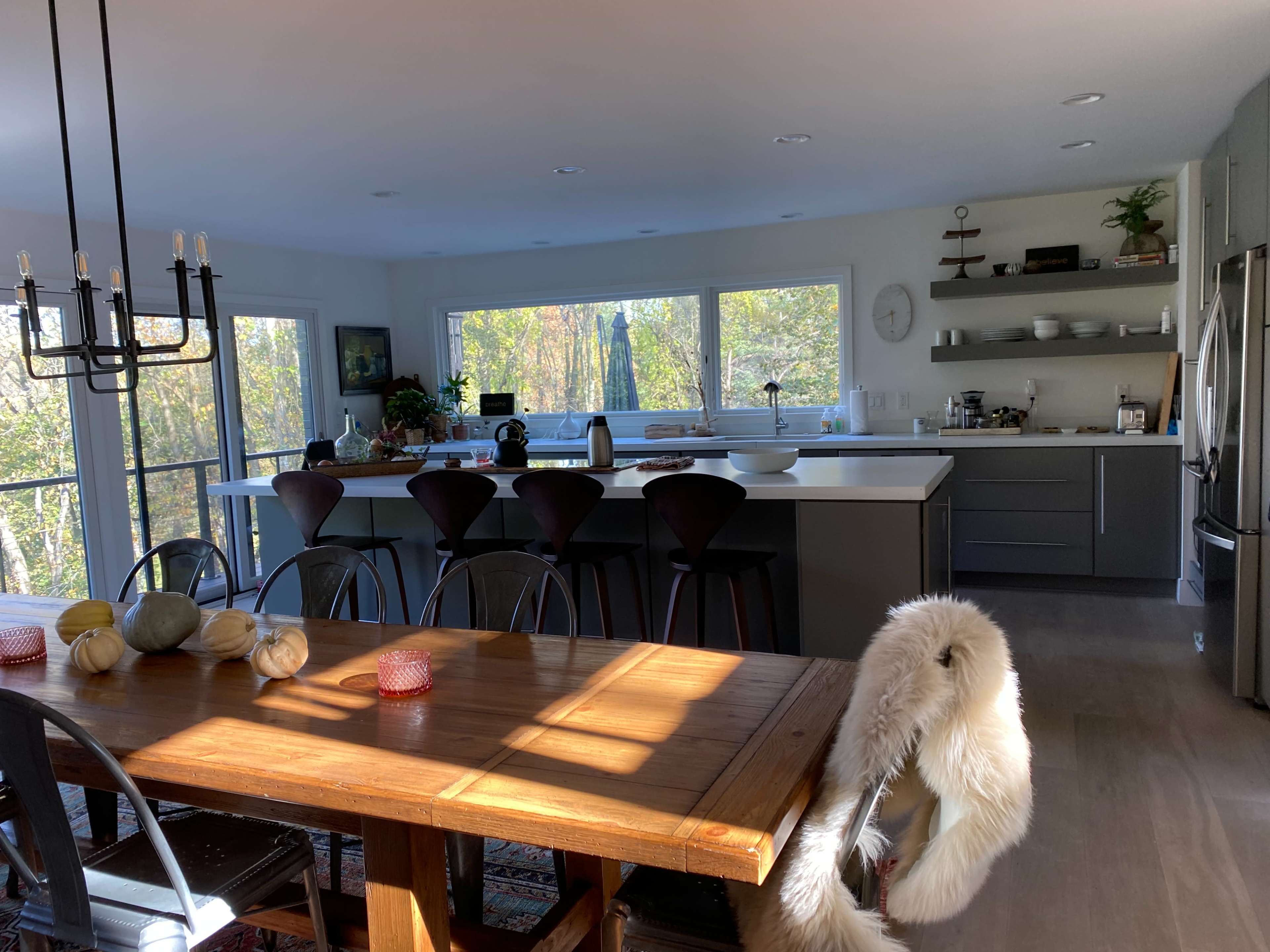 A modern kitchen with a large island and a dining table, featuring large windows that offer a view of greenery outside.