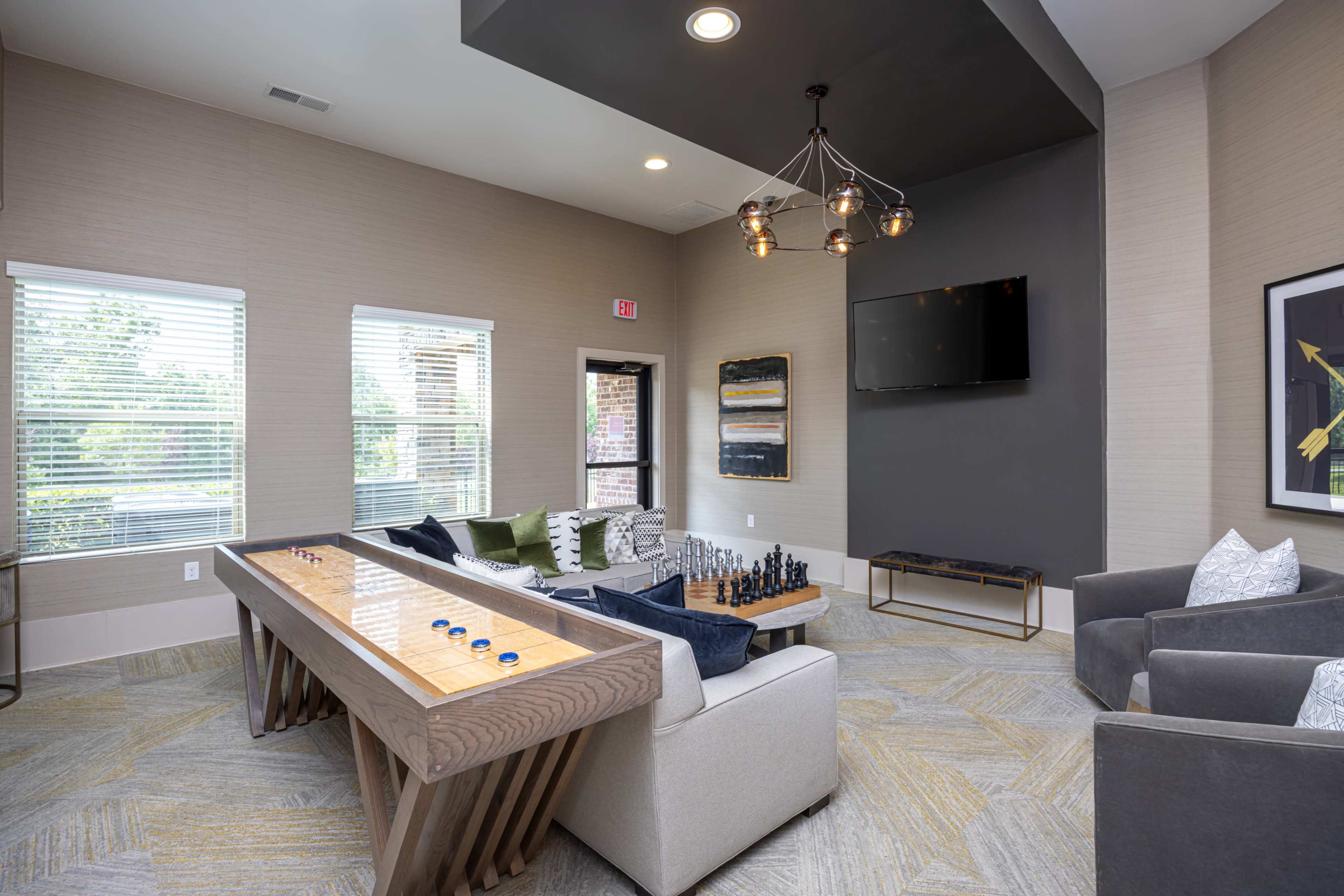 The image shows a modern lounge area with a large wooden game table, a seating arrangement including a sofa and armchairs, and a wall-mounted television.