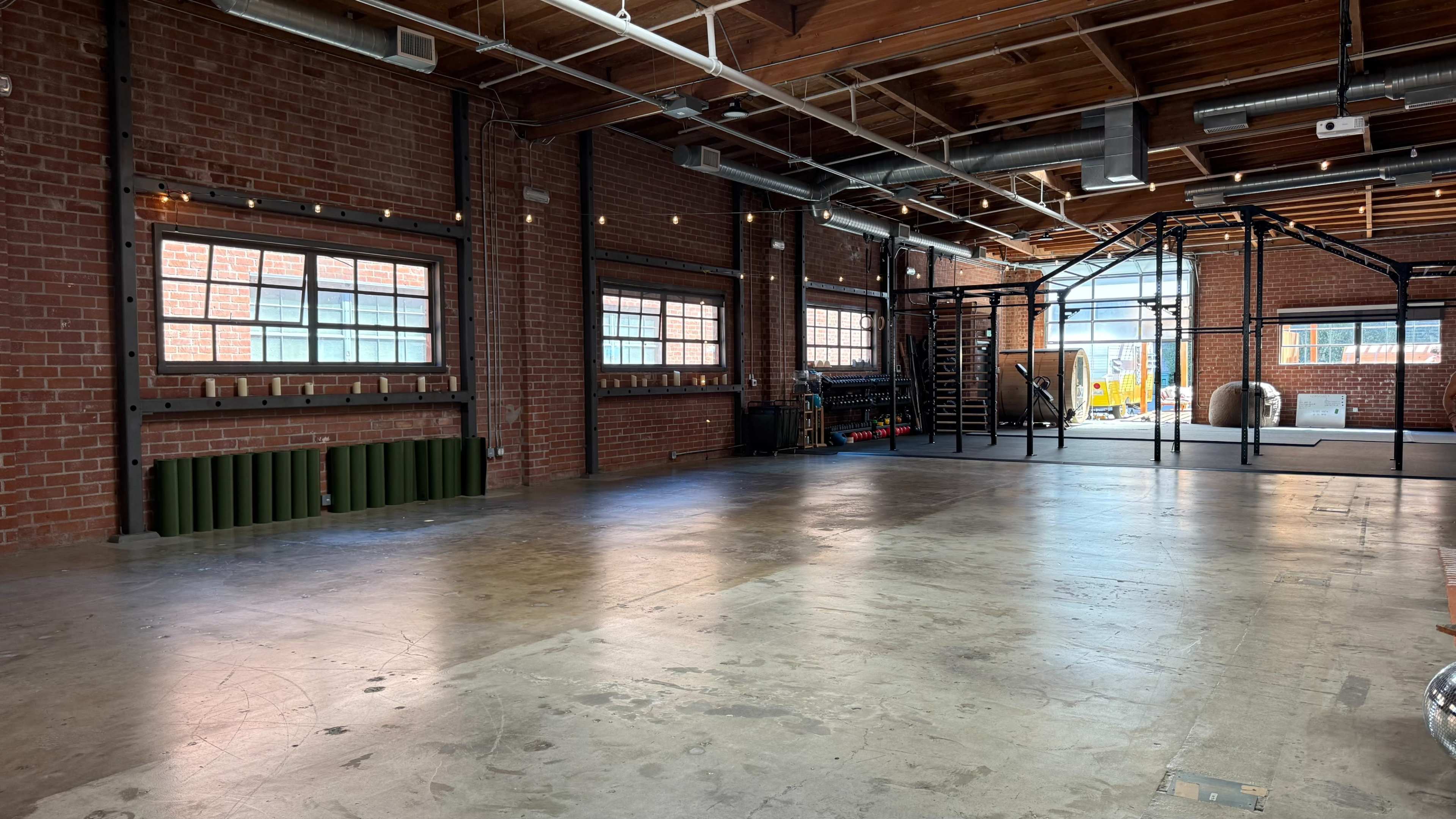 The image shows a spacious gym with exposed brick walls, large windows, and open areas for workout equipment and exercise activities.