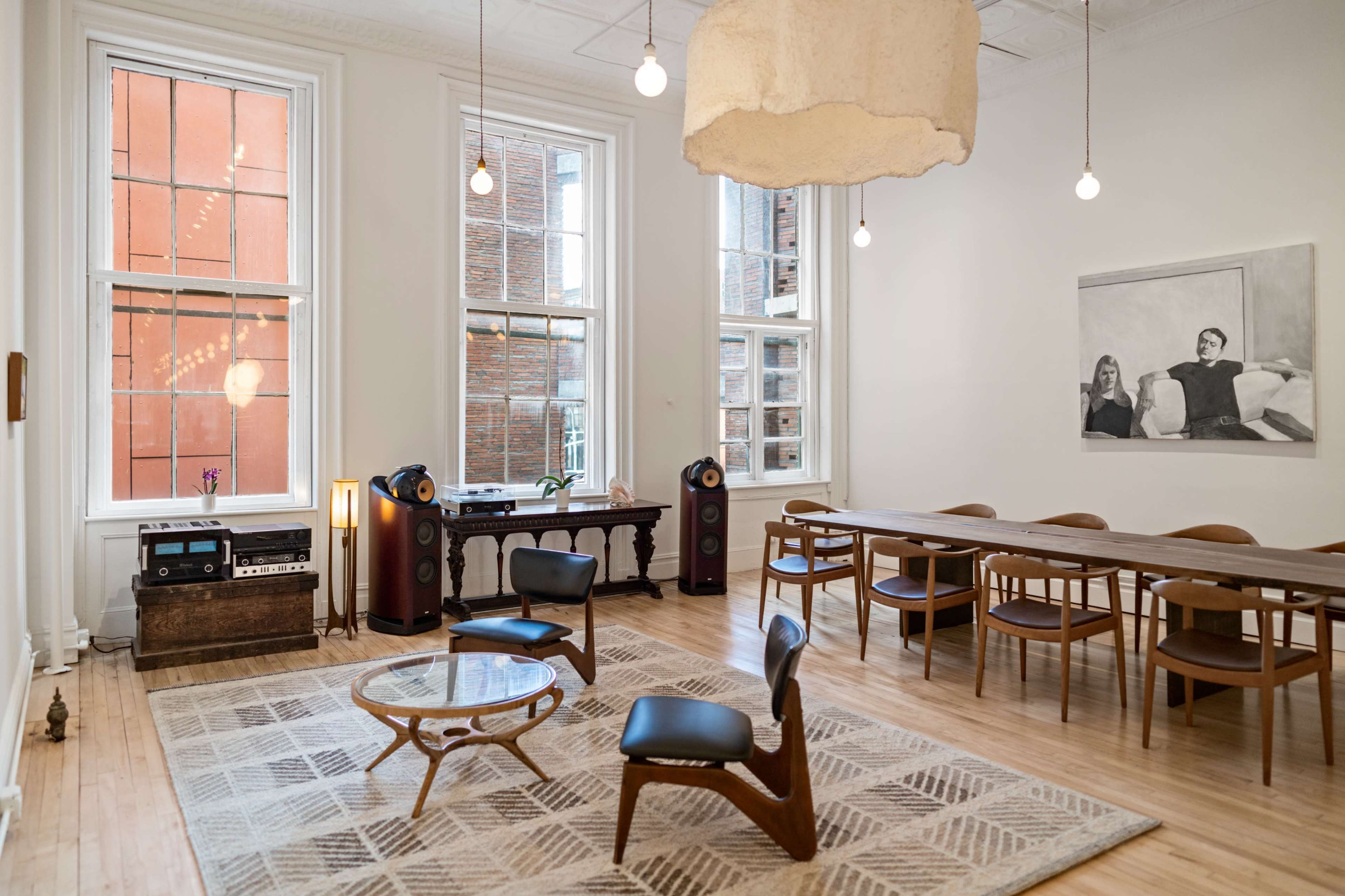 A spacious room with large windows, wooden floors, a dining table, and modern furniture including a lounge chair and audio equipment.