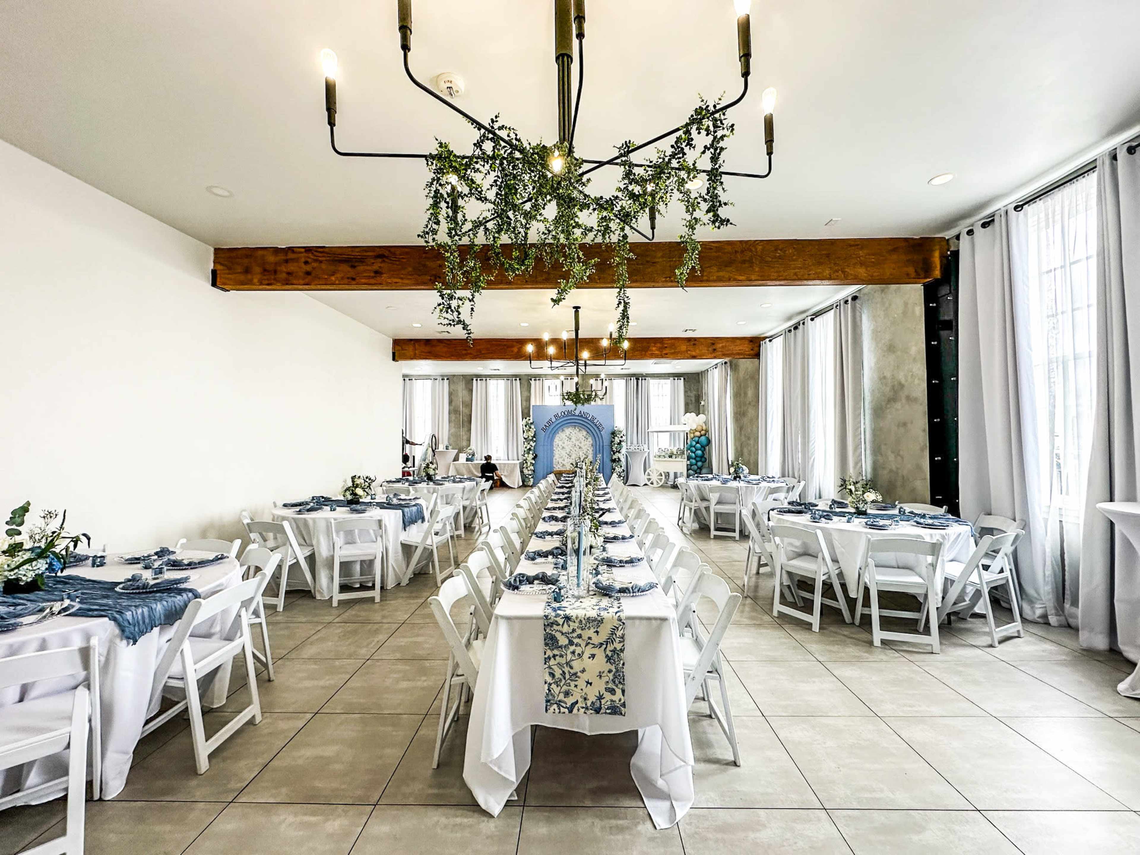 The image shows a spacious dining area set up with long tables adorned with white tablecloths and blue runners, surrounded by foliage and soft lighting fixtures.