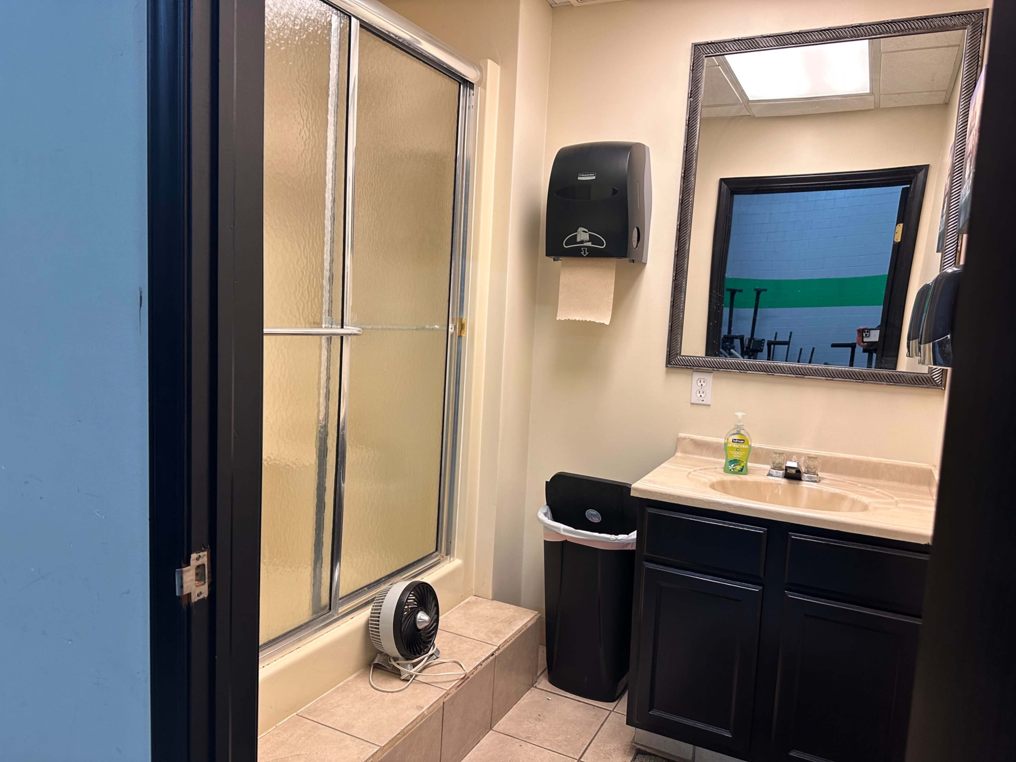 A bathroom with a shower stall, a sink with a mirror above it, a trash can, a hand dryer, and a floor fan.