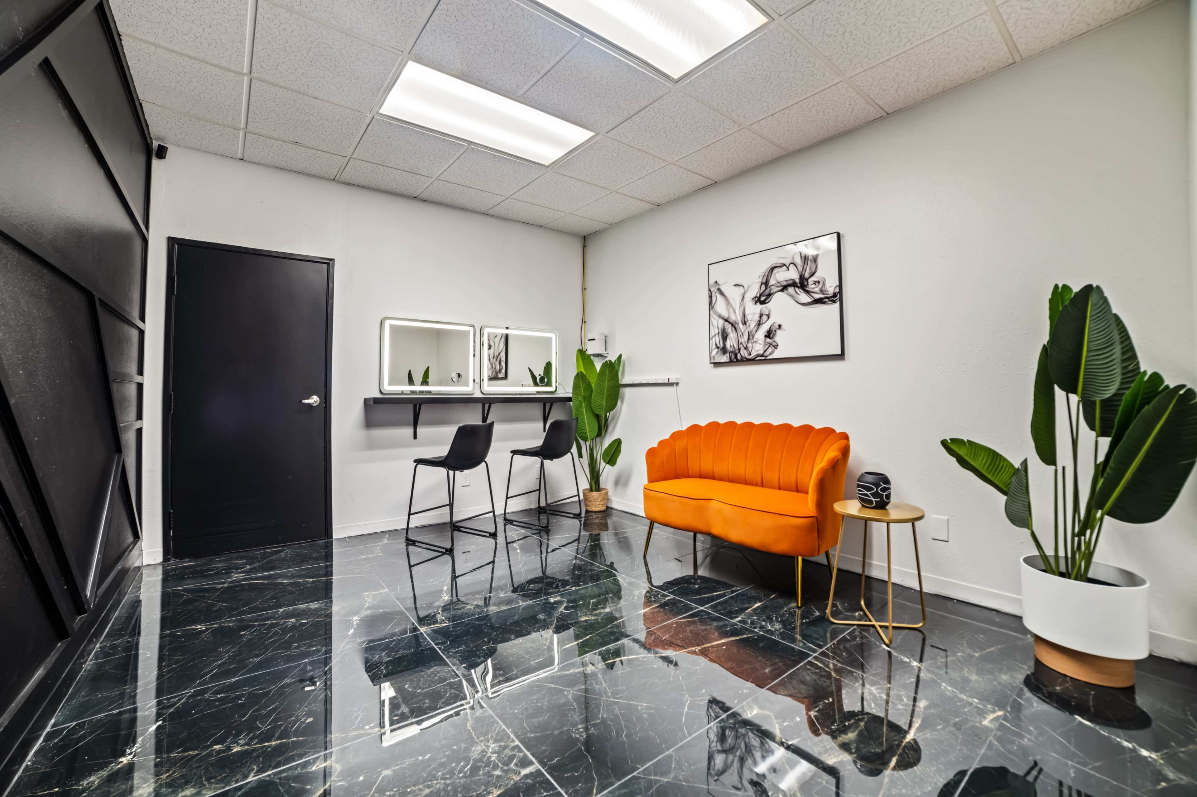 A modern interior space featuring an orange couch, two black chairs, a wall-mounted mirror, and large potted plants on a glossy black floor.