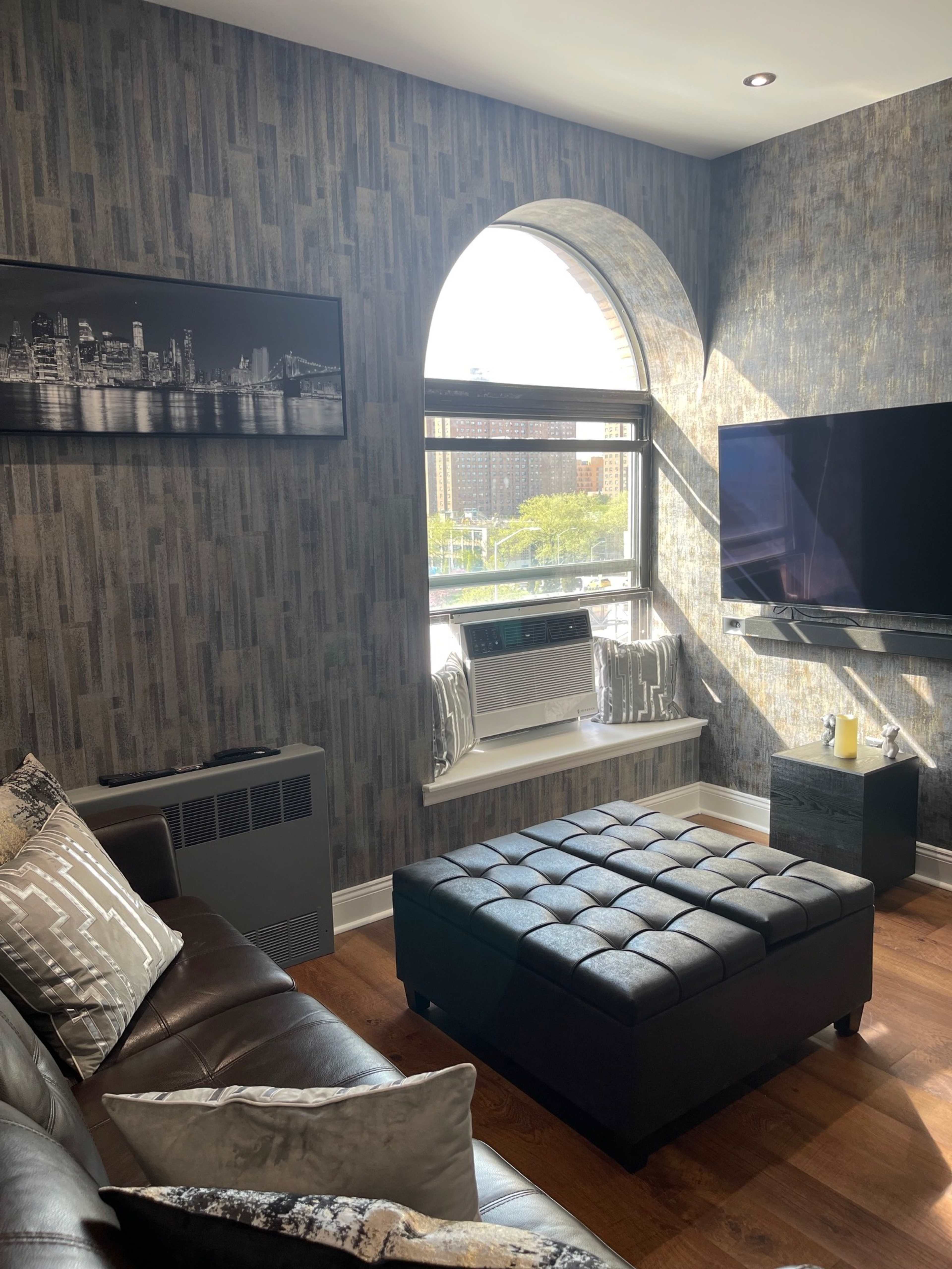 A cozy living room features a brown leather sofa, a black tufted ottoman, and a wall-mounted television beneath a large arched window overlooking an urban view.