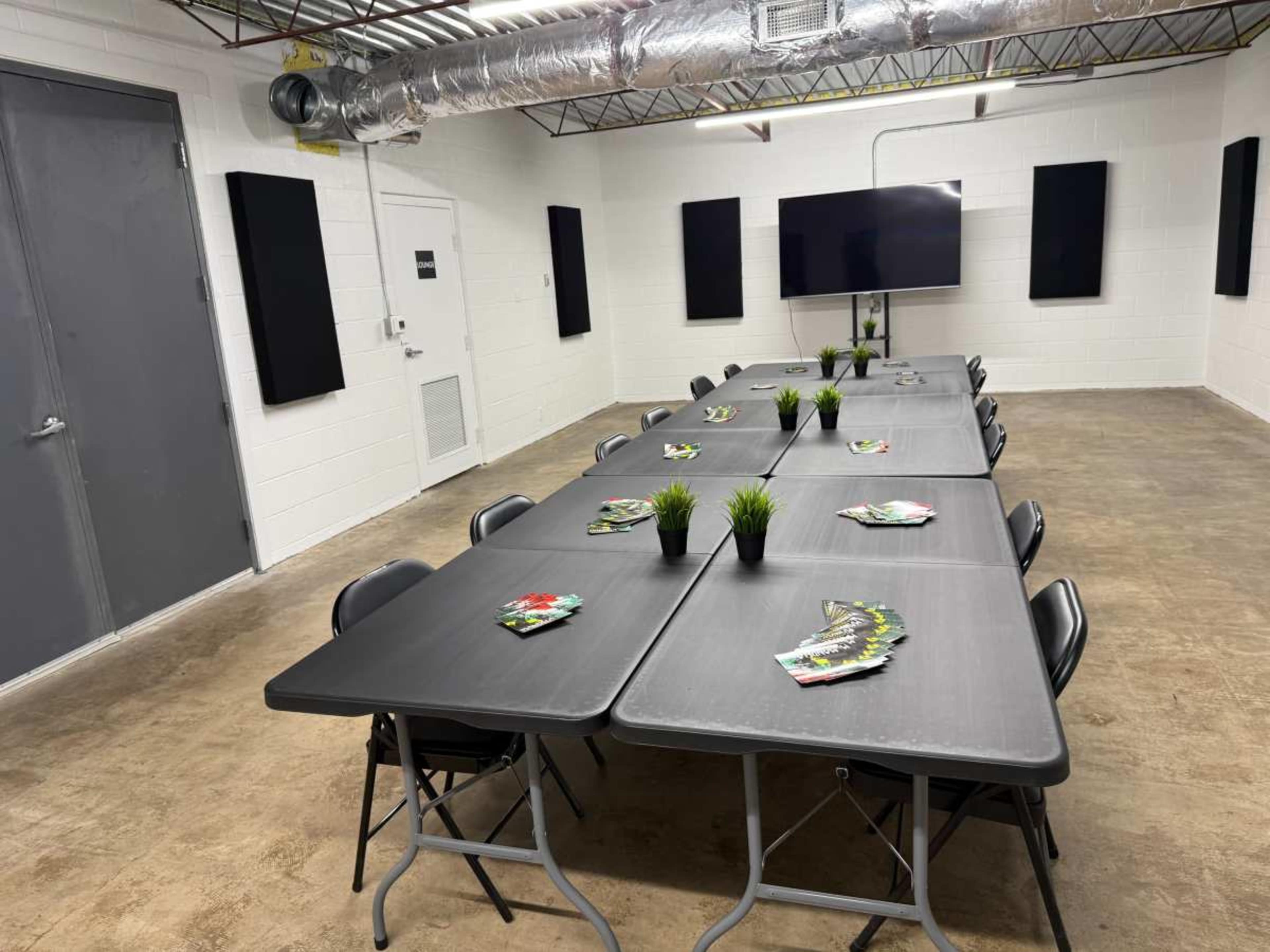 A long rectangular table is set up in a sparsely decorated room, with black folding chairs on either side and small potted plants and pamphlets placed on the table.