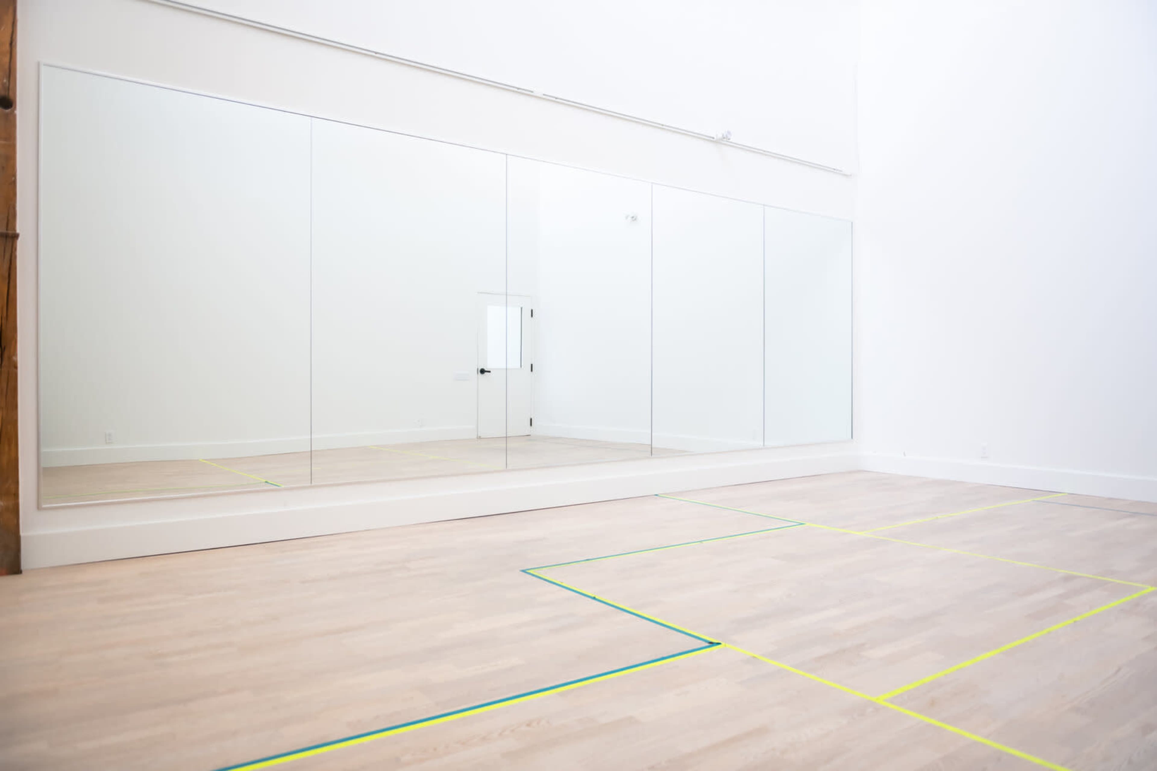 A bright, empty room with large mirrors on one wall and a wooden floor marked by yellow and green lines.