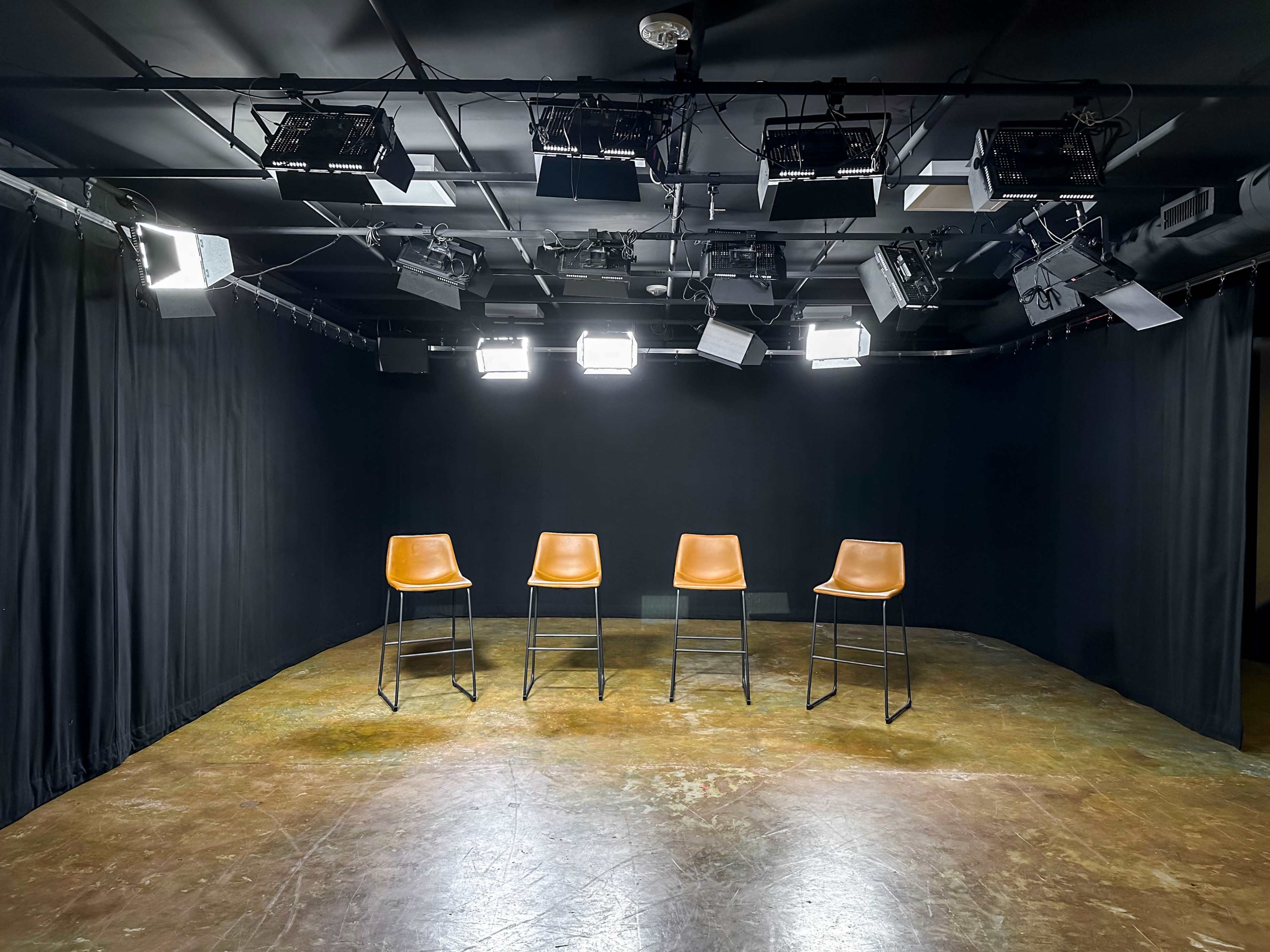 A studio setup with four empty bar stools arranged in a row under bright stage lights against a black backdrop.