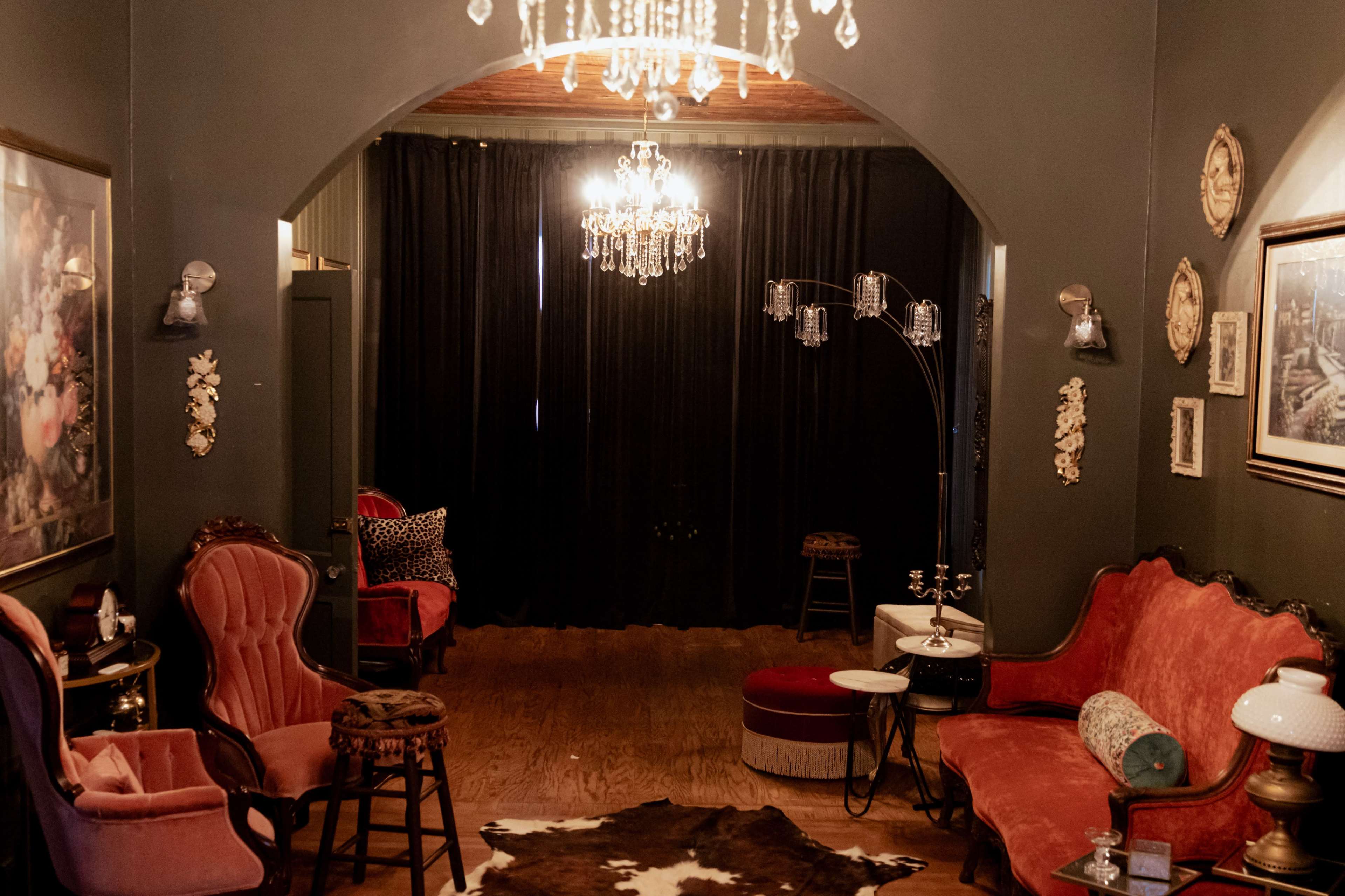 The image shows a stylishly decorated room featuring vintage furniture, including a red couch and armchairs, with chandeliers hanging from the ceiling and dark curtains in the background.