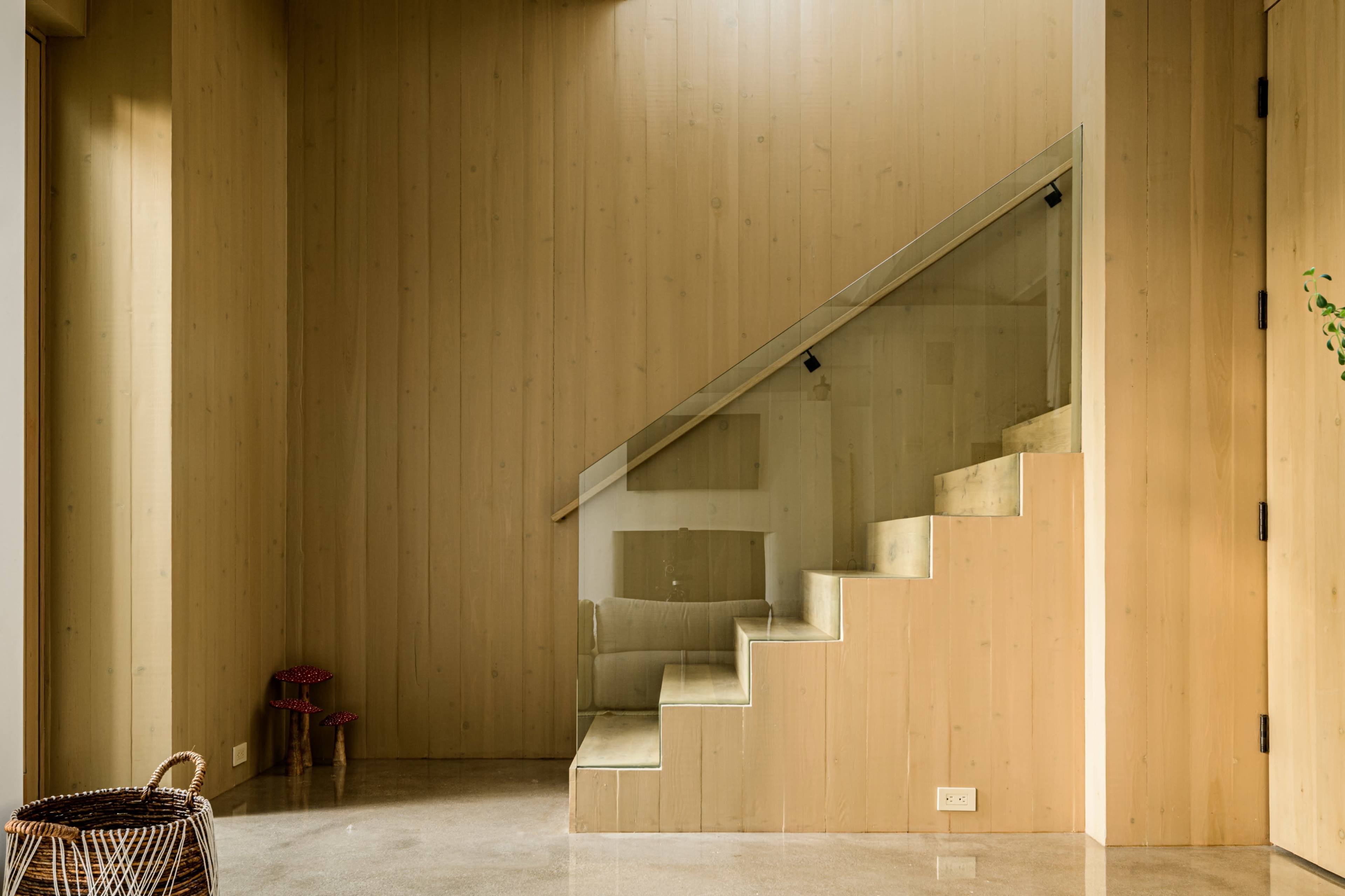 A wooden interior with a staircase leading to an upper level, flanked by a glass railing and a basket in the corner.
