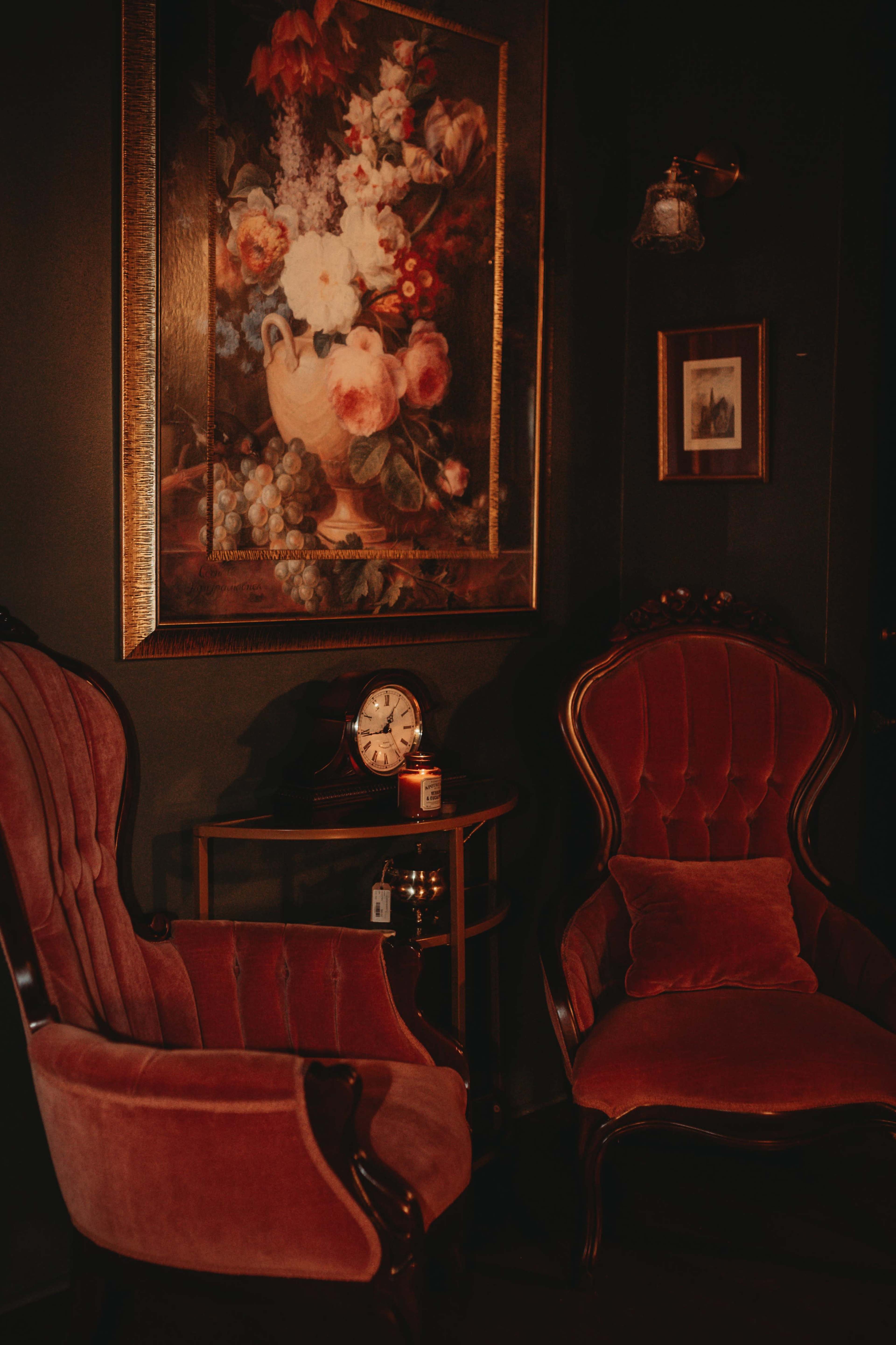The image features two vintage velvet chairs beside a small table with a clock, all placed against a dark wall adorned with a floral painting.